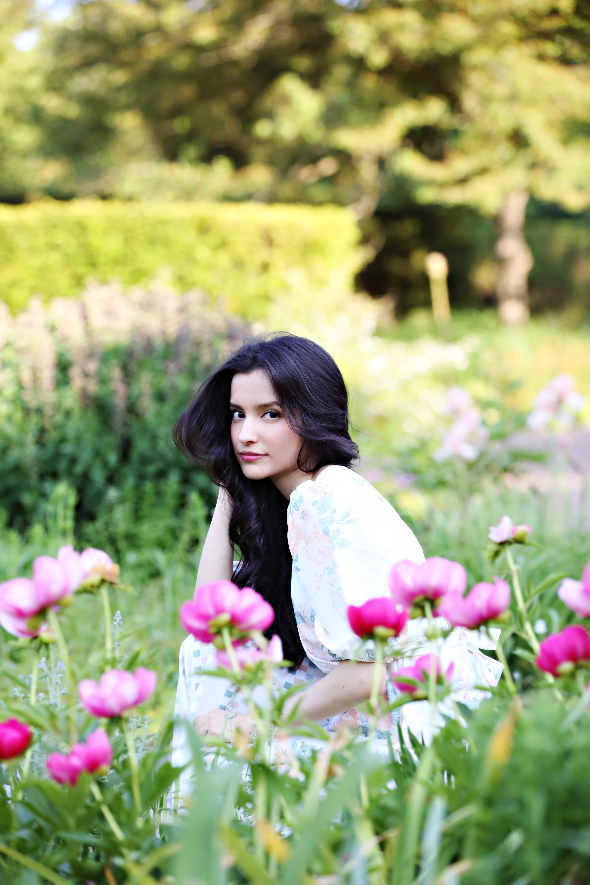 A young woman with long dark hair sitting among pink flowers in a lush green garden.