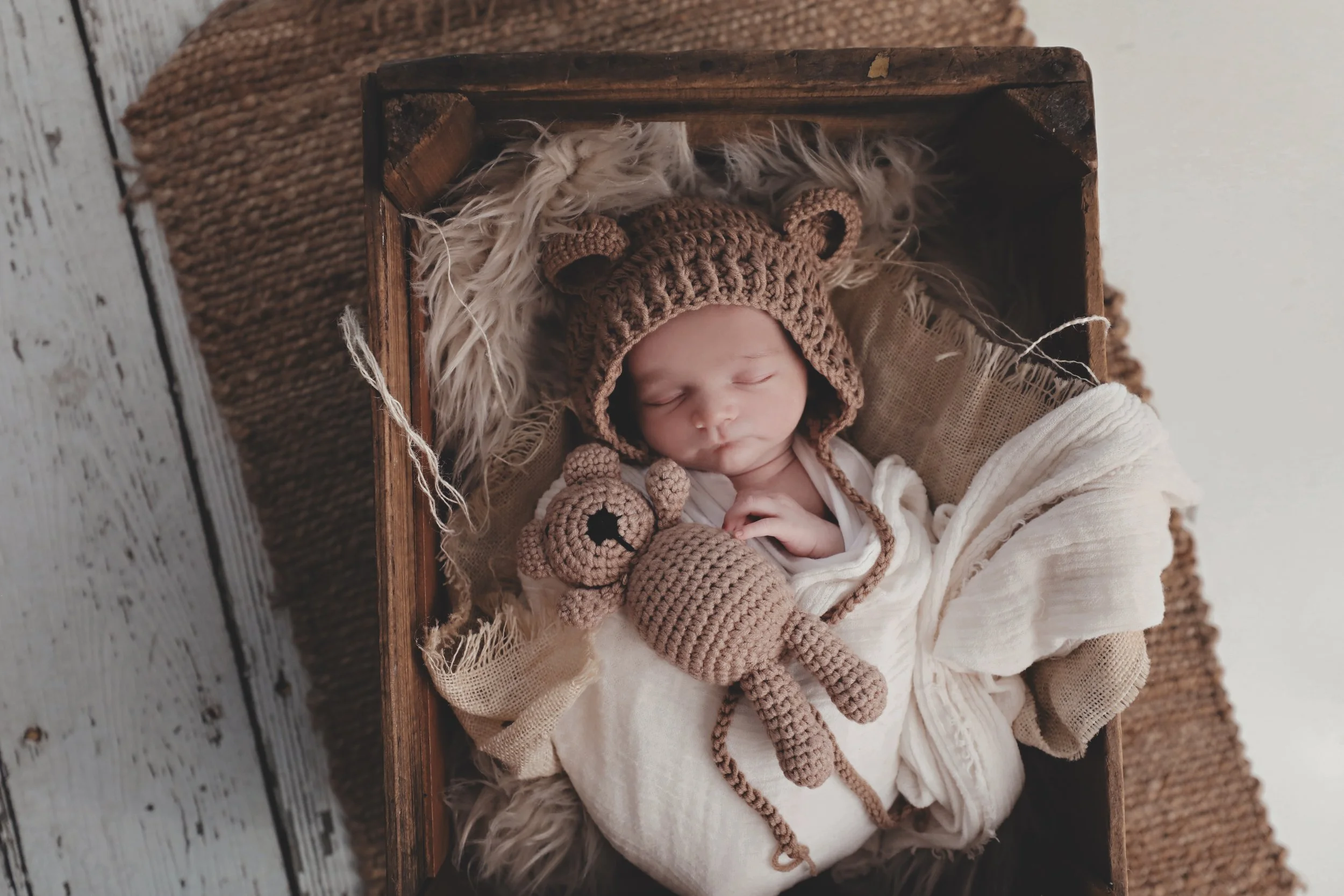 A newborn baby sleeping in a wooden basket, wearing a brown crochet bear hat. The baby holds a small crochet bear toy, and the basket is lined with soft fabric and fur. The background includes a woven mat and a wooden surface.