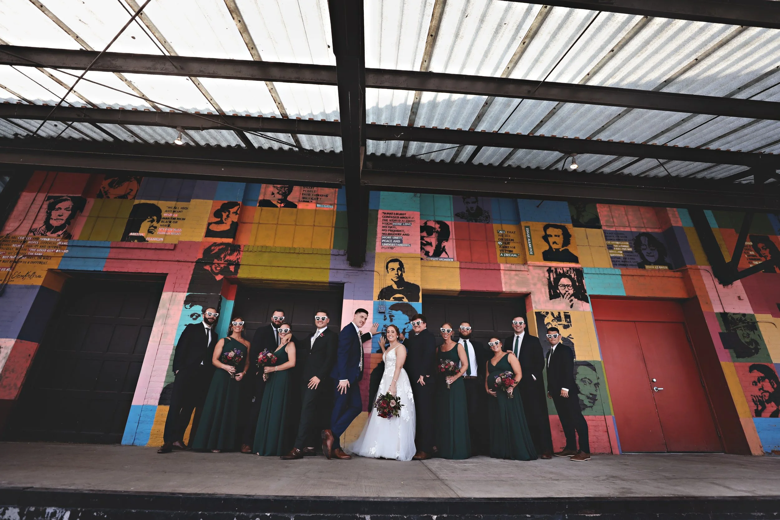 A wedding party standing in front of a colorful mural with portraits and quotes, with the bride in a white gown holding a bouquet and the groom in a suit, surrounded by bridesmaids and groomsmen wearing matching outfits, all wearing sunglasses.