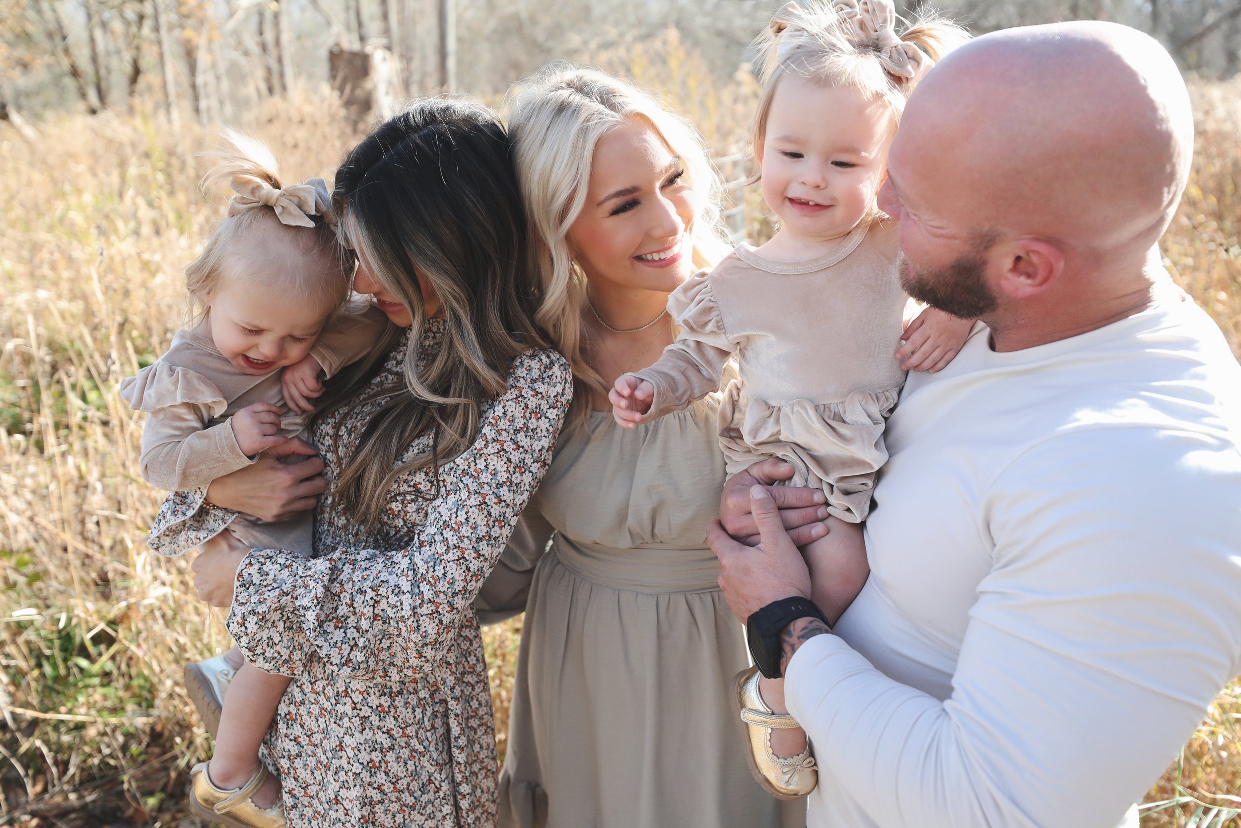 A family of five, including two women, one man, and two young girls, smiling and enjoying time together outdoors in a field with tall grass and trees, during a sunny day.