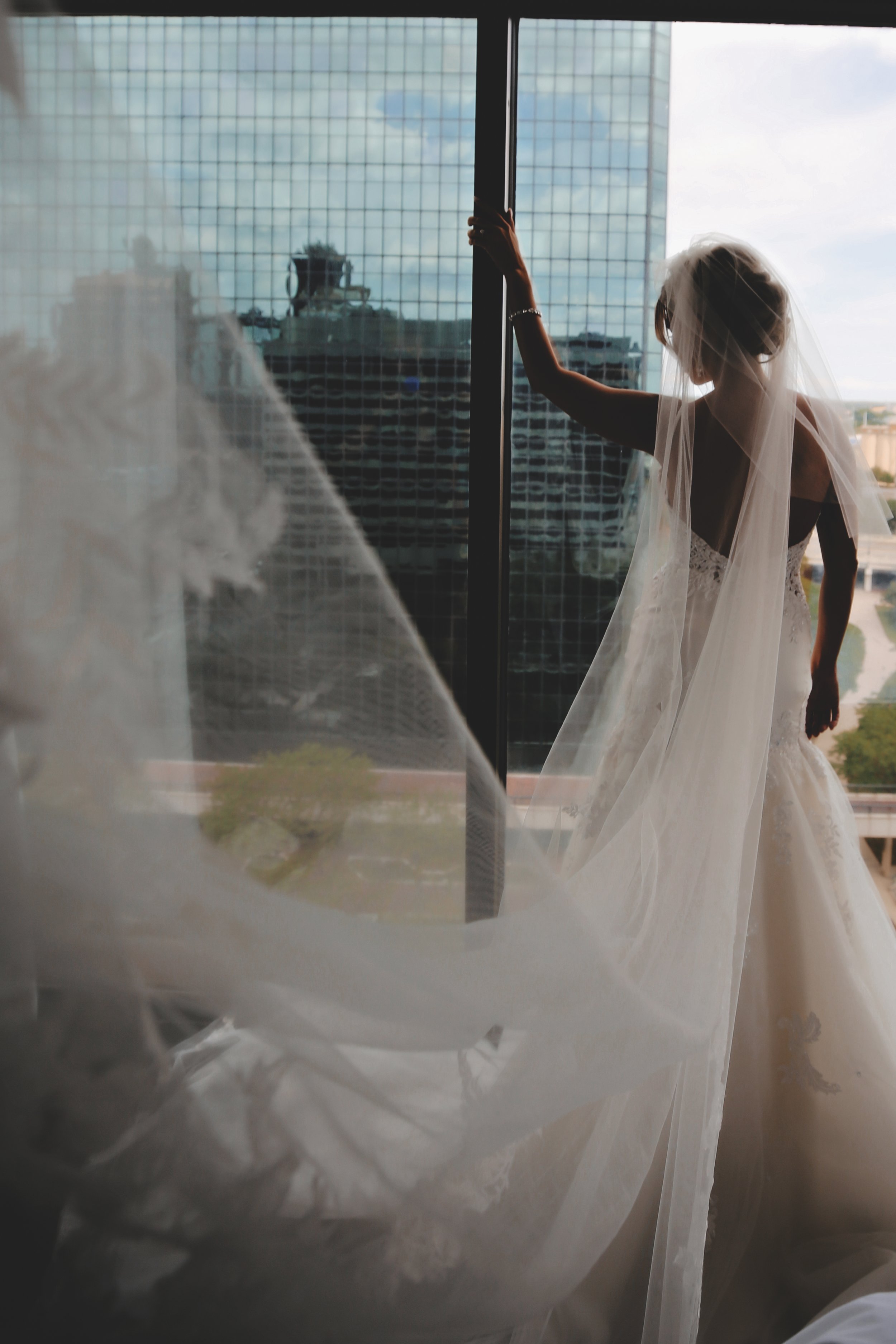 A bride in a wedding dress standing near a large window, with her hand on the window frame, gazing outside. The window reflects the cityscape with tall buildings.