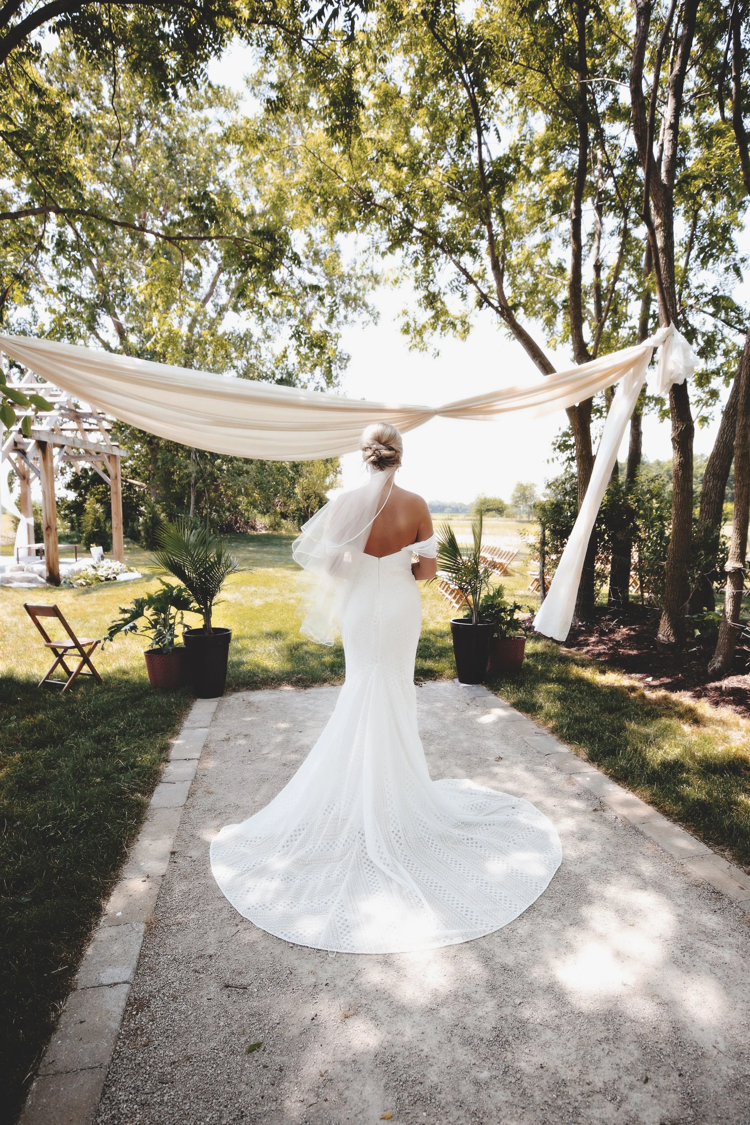 Bride in a white wedding gown walking away under a draped white fabric arch outdoors, surrounded by trees and potted plants.