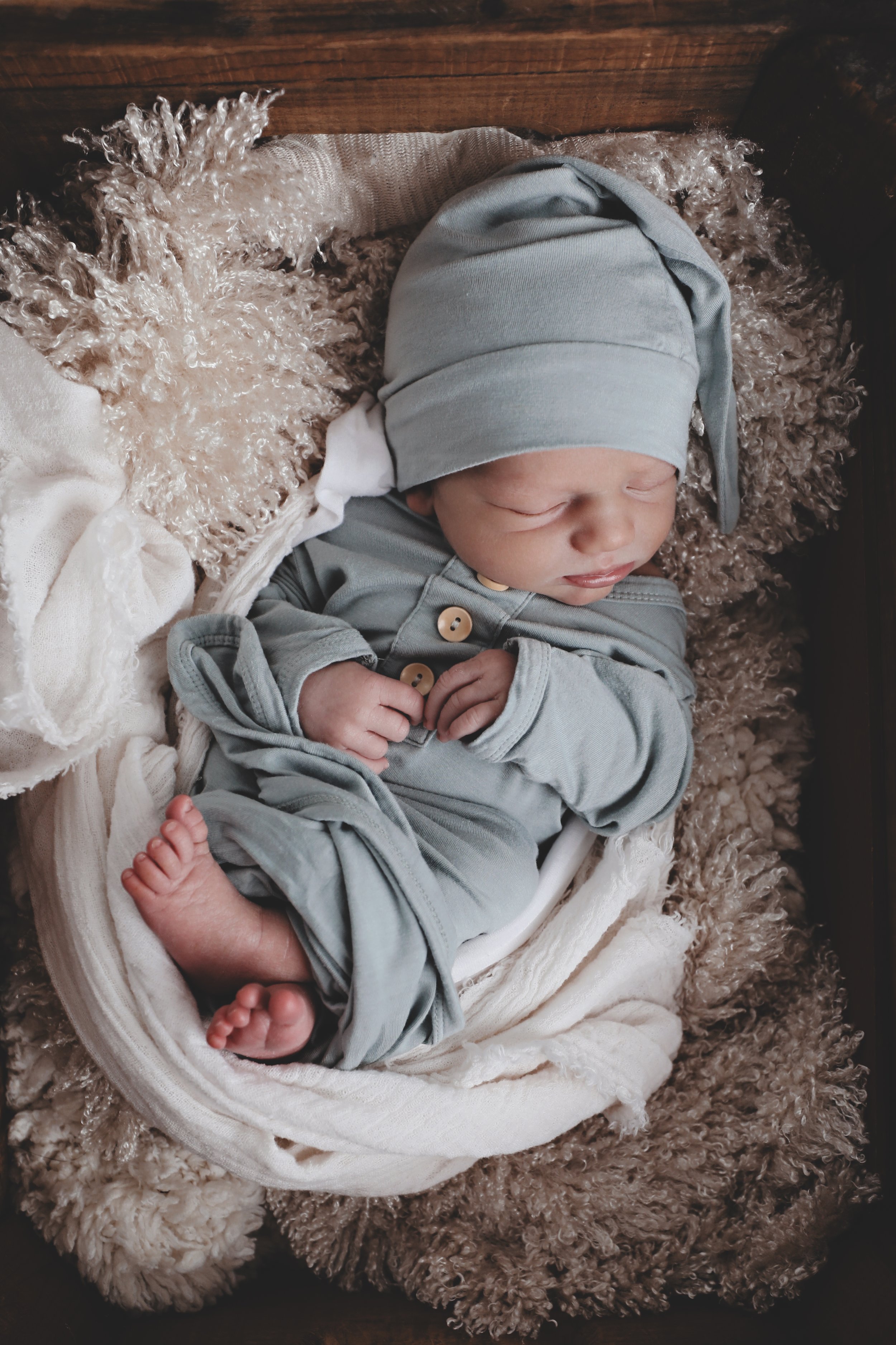 A sleeping baby wearing a gray hat and matching gray pajamas, resting on a soft, textured blanket surrounded by fluffy, curly fabric.