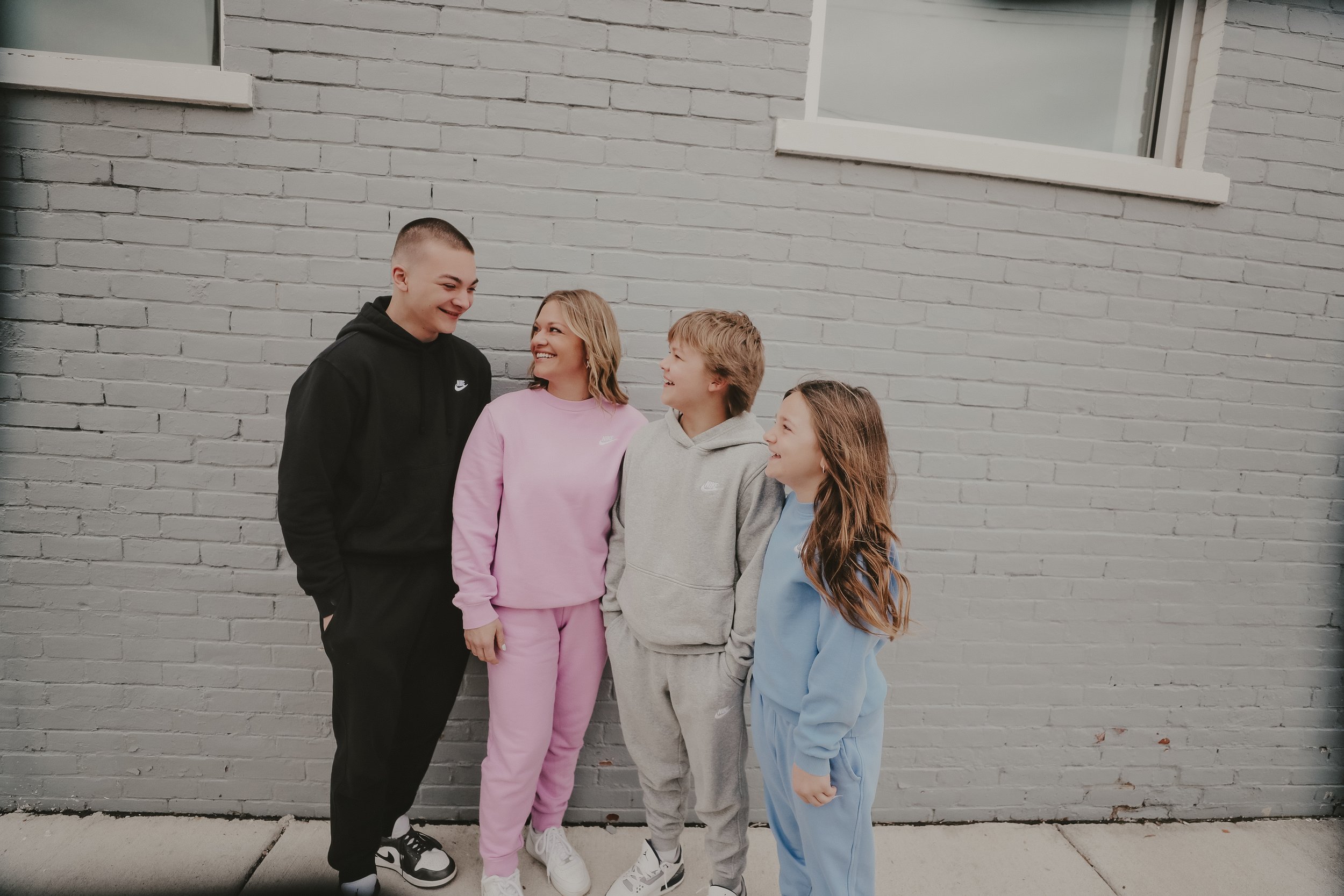 Group of five young people standing against a light gray brick wall, smiling and enjoying each other's company, dressed in colorful Nike hoodies and sweatpants.