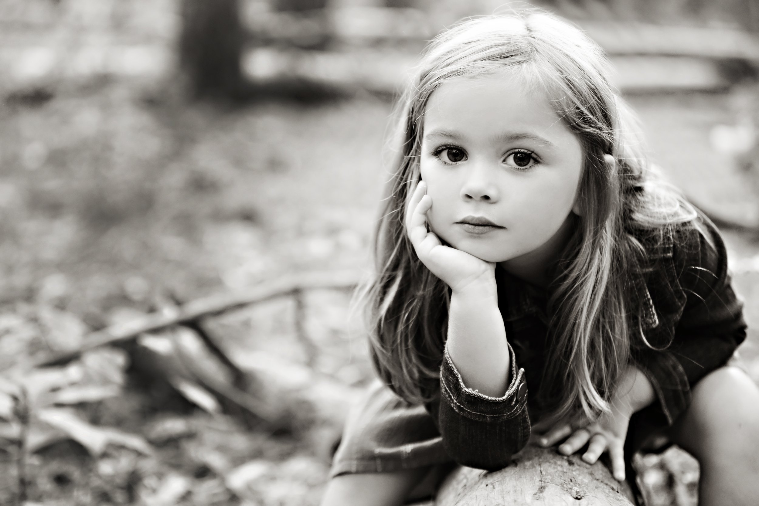 A black and white photo of a young girl with long hair, resting her face on her hand while crouching on the ground outdoors.