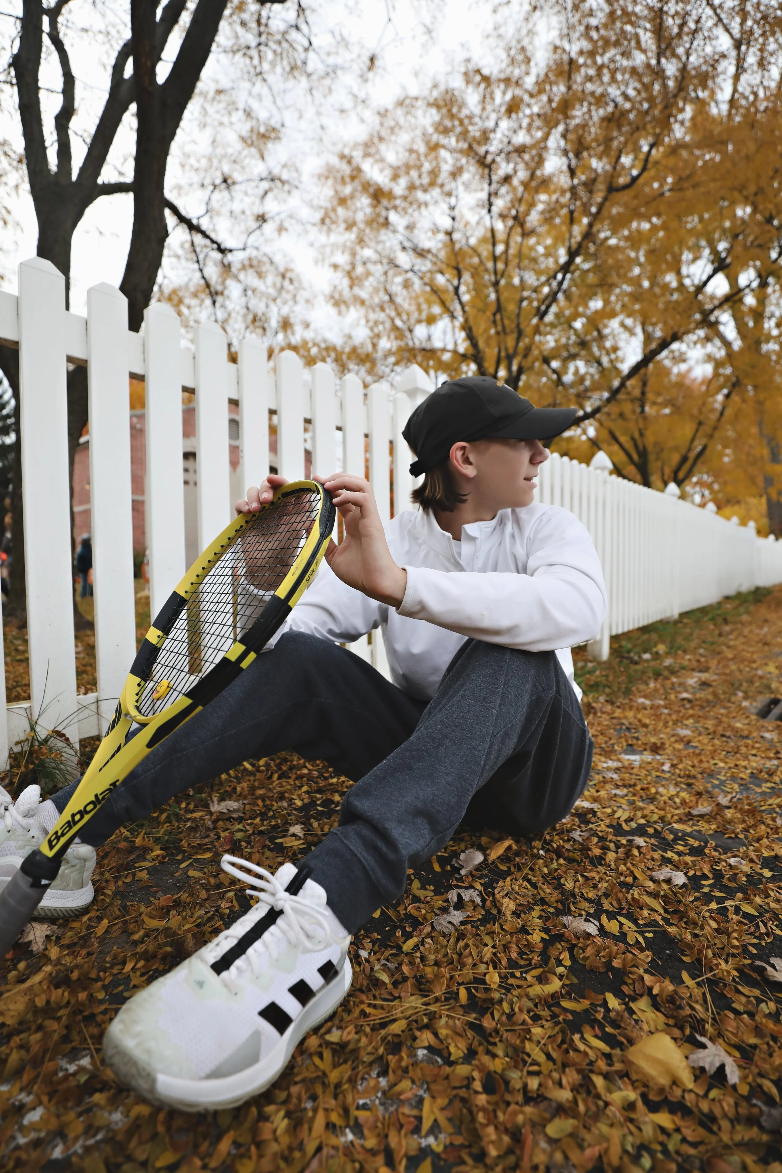 Young person sitting on the ground outdoors during autumn, holding a tennis racket, wearing a black cap, white jacket, black pants, and white sneakers, next to a white picket fence and fallen leaves, with trees with orange leaves in the background.