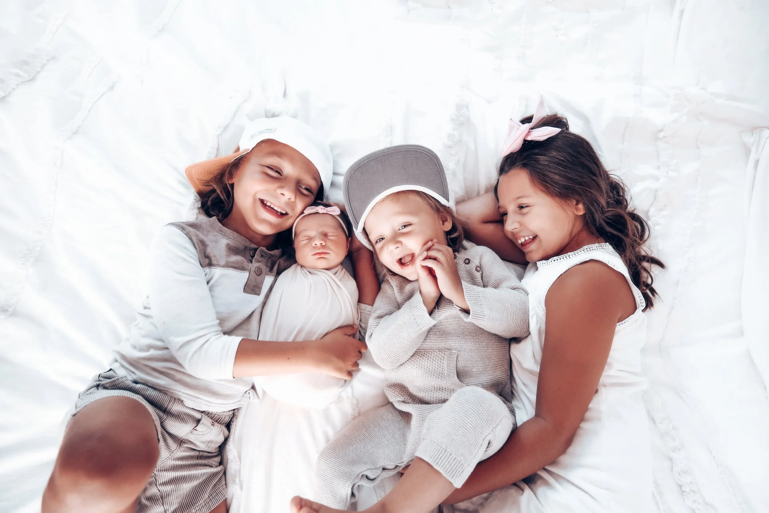 Four children lying down on white bedding, smiling and cuddling each other, with a baby sleeping in the middle.