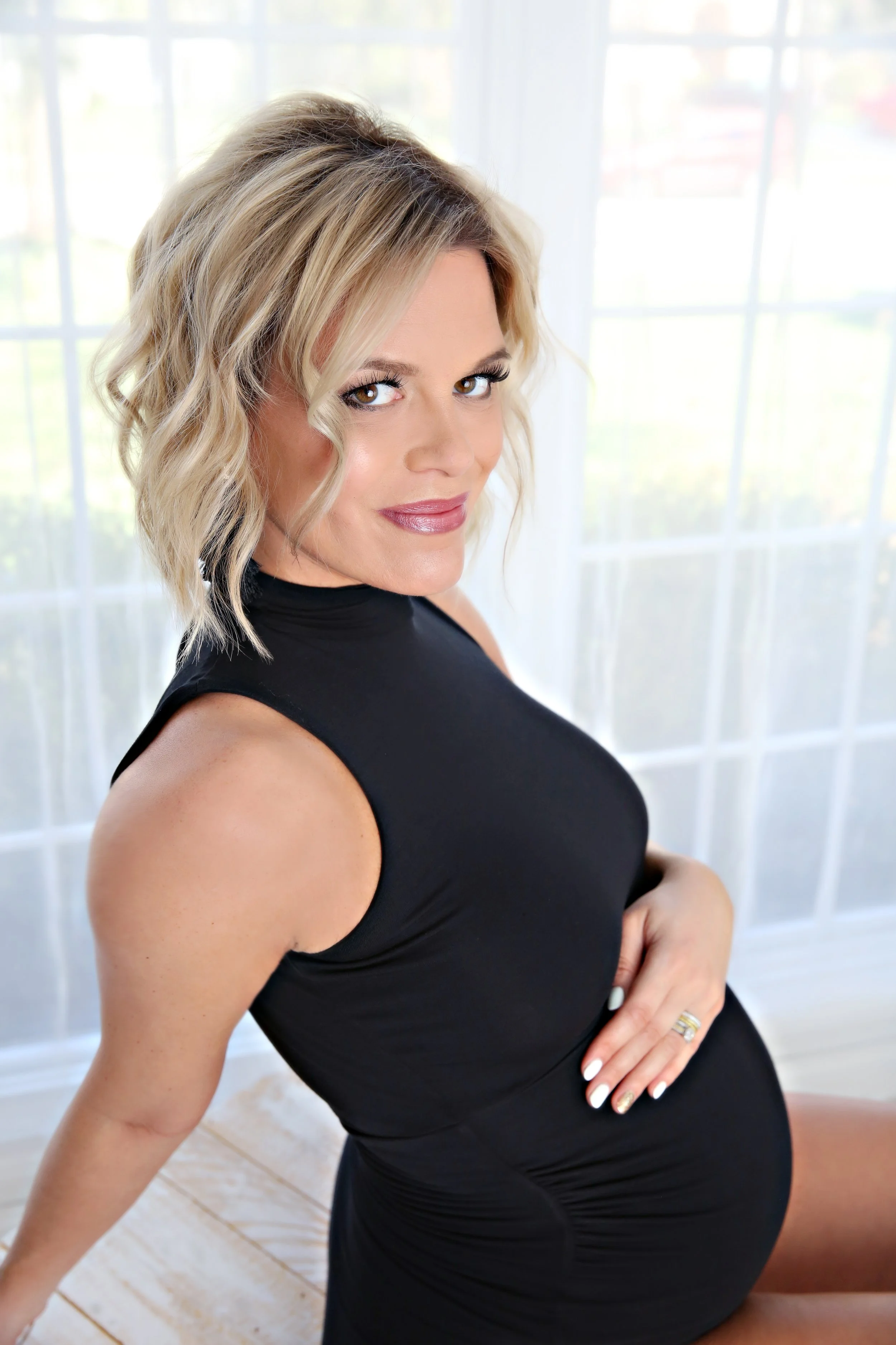 A pregnant woman with shoulder-length blonde hair styled in loose waves, wearing a sleeveless black dress, sitting indoors near a large window with sheer curtains, smiling and looking at the camera.
