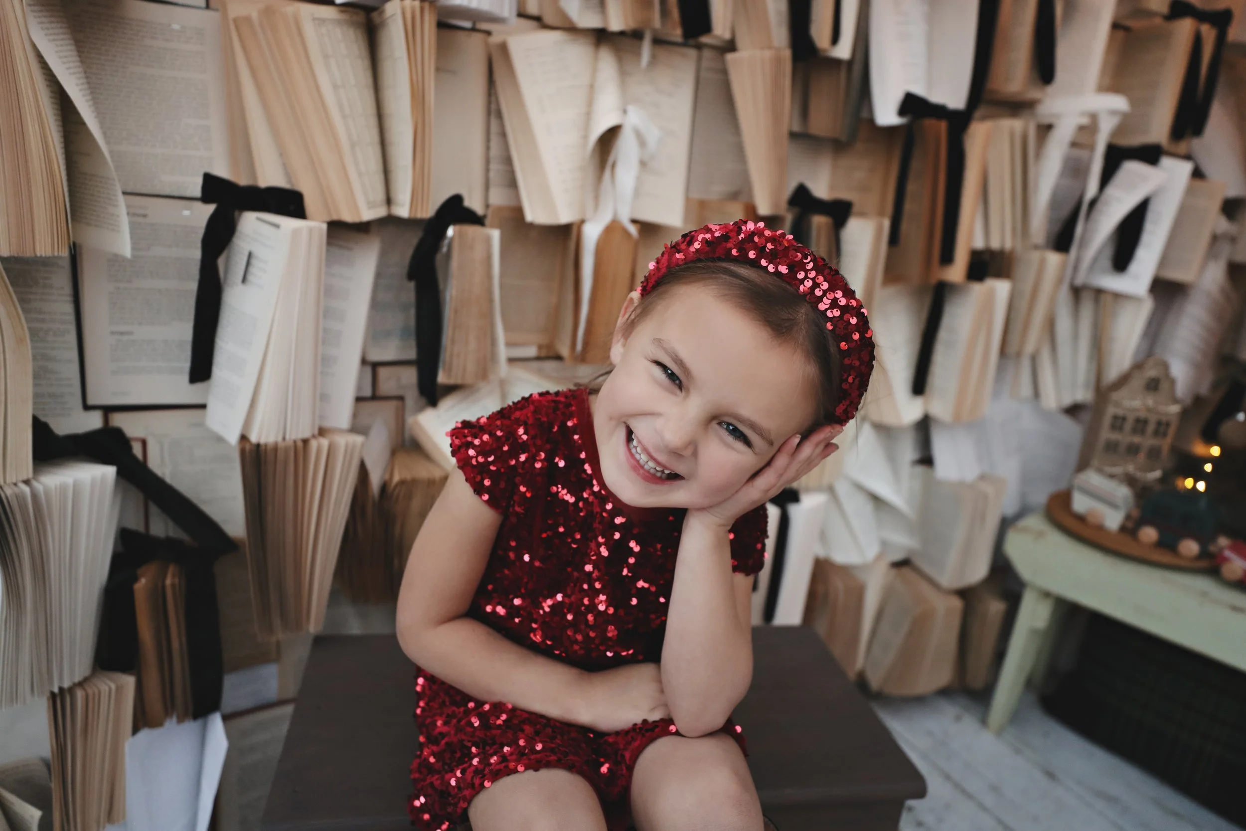 A young girl in a red sequined dress and matching headband sitting on a bench, smiling and leaning her head on her hand, with a wall of open and closed books behind her.