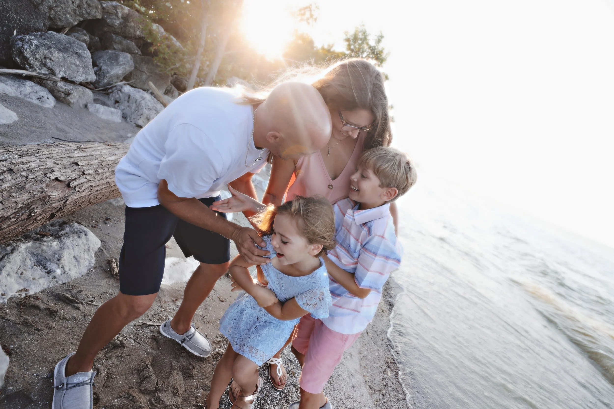 A family of four enjoying a moment together on the beach during sunset, with a rocky shoreline behind them and water on the right side.