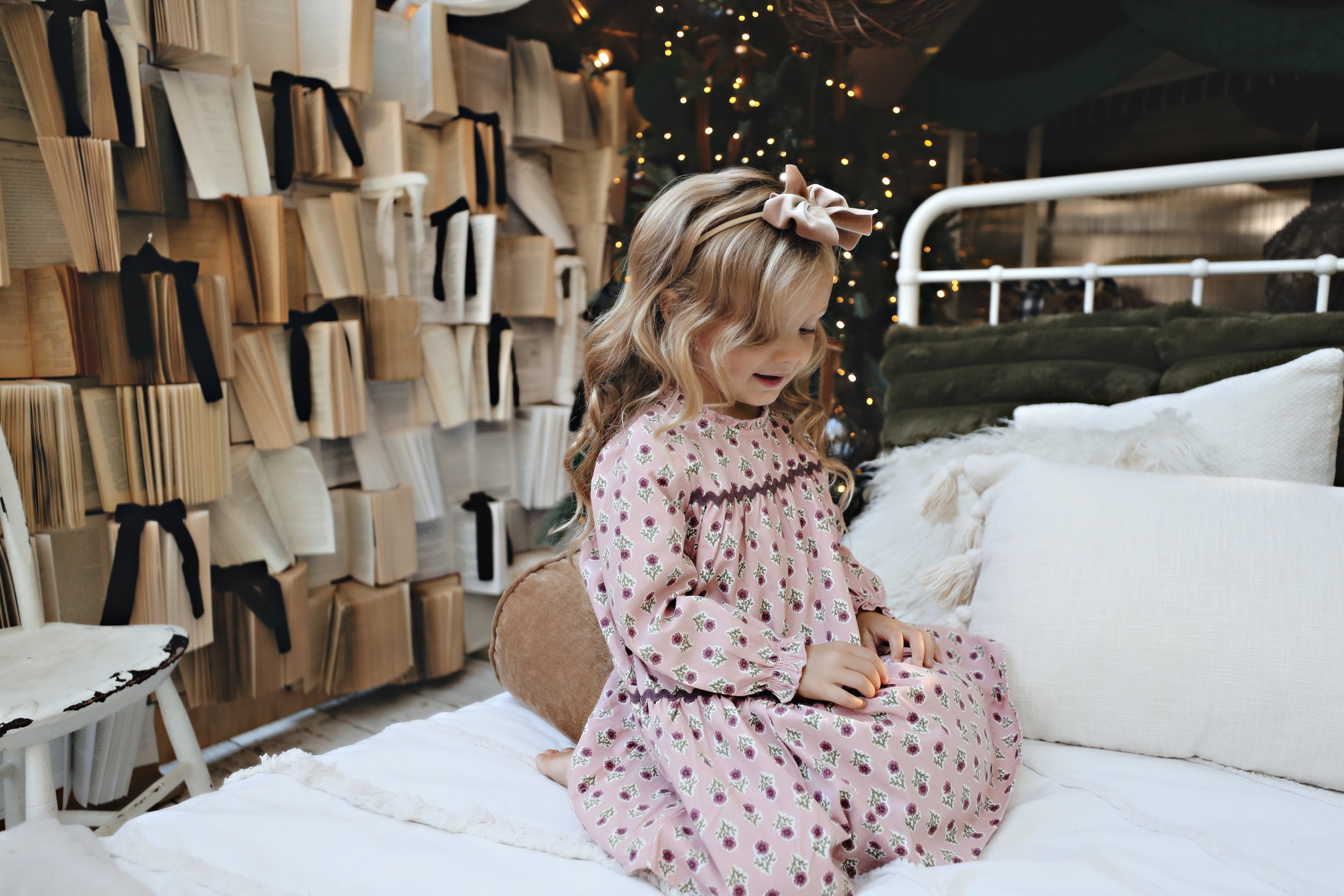 A young girl with long curly hair and a pink bow sitting on a white bed in a cozy, decorated room with books on the wall and Christmas lights in the background.