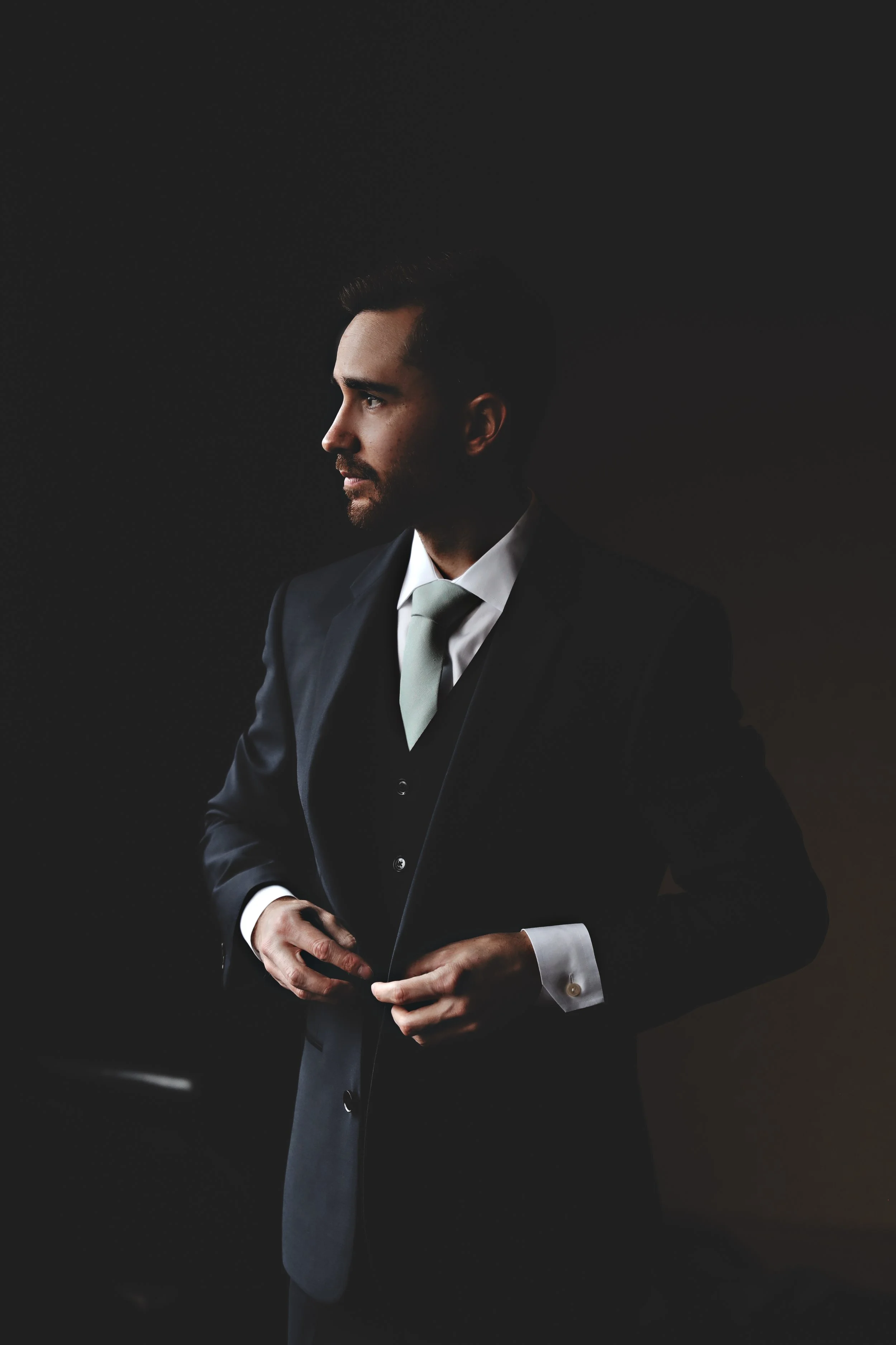 A man in a dark suit, white shirt, and light-colored tie adjusting his jacket, standing against a dark background.