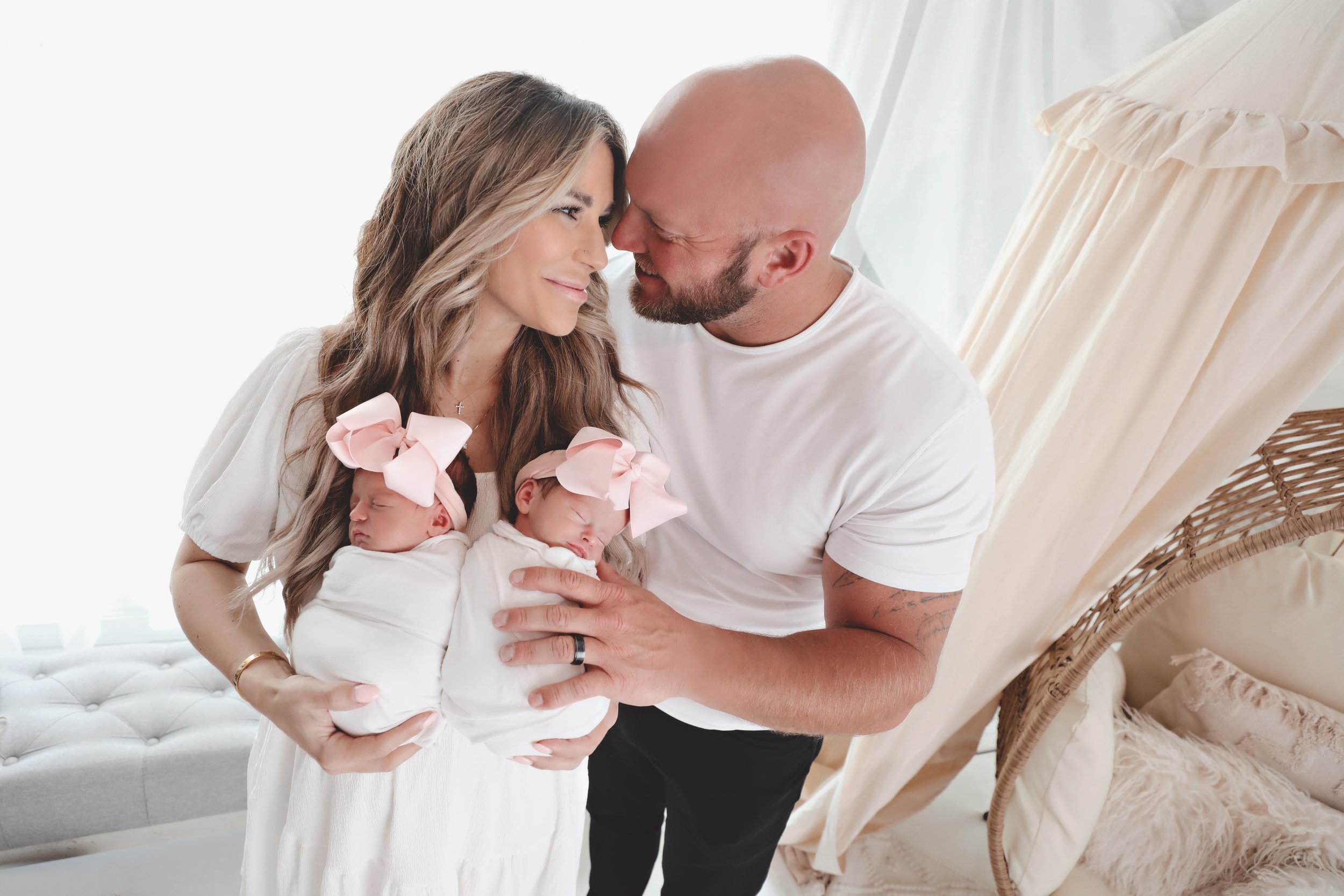 A family with a mother, father, and two newborn babies with pink bows in a cozy, light-colored nursery.