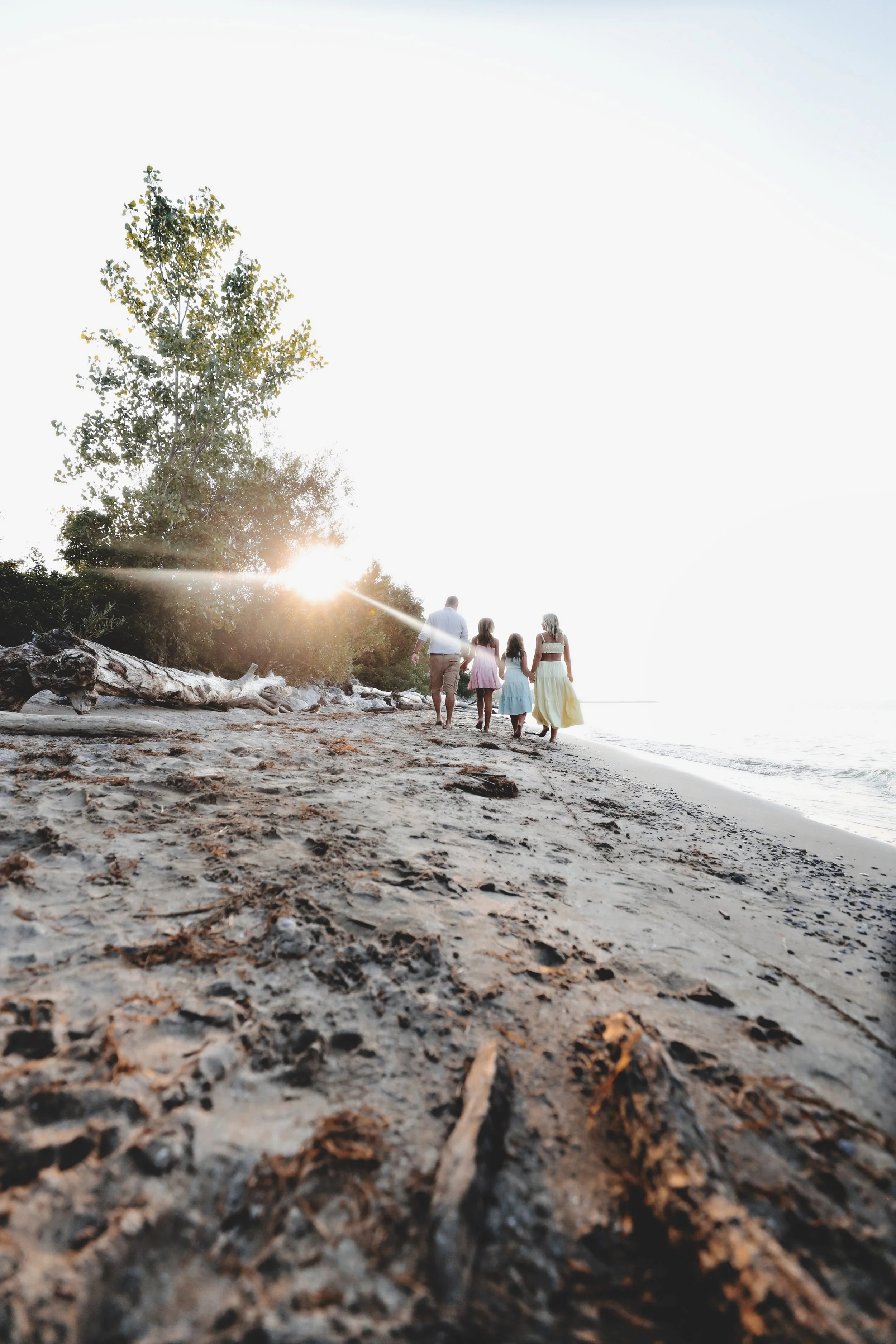A family of four walking along a sandy beach during sunset, with trees in the background and the sun low on the horizon.