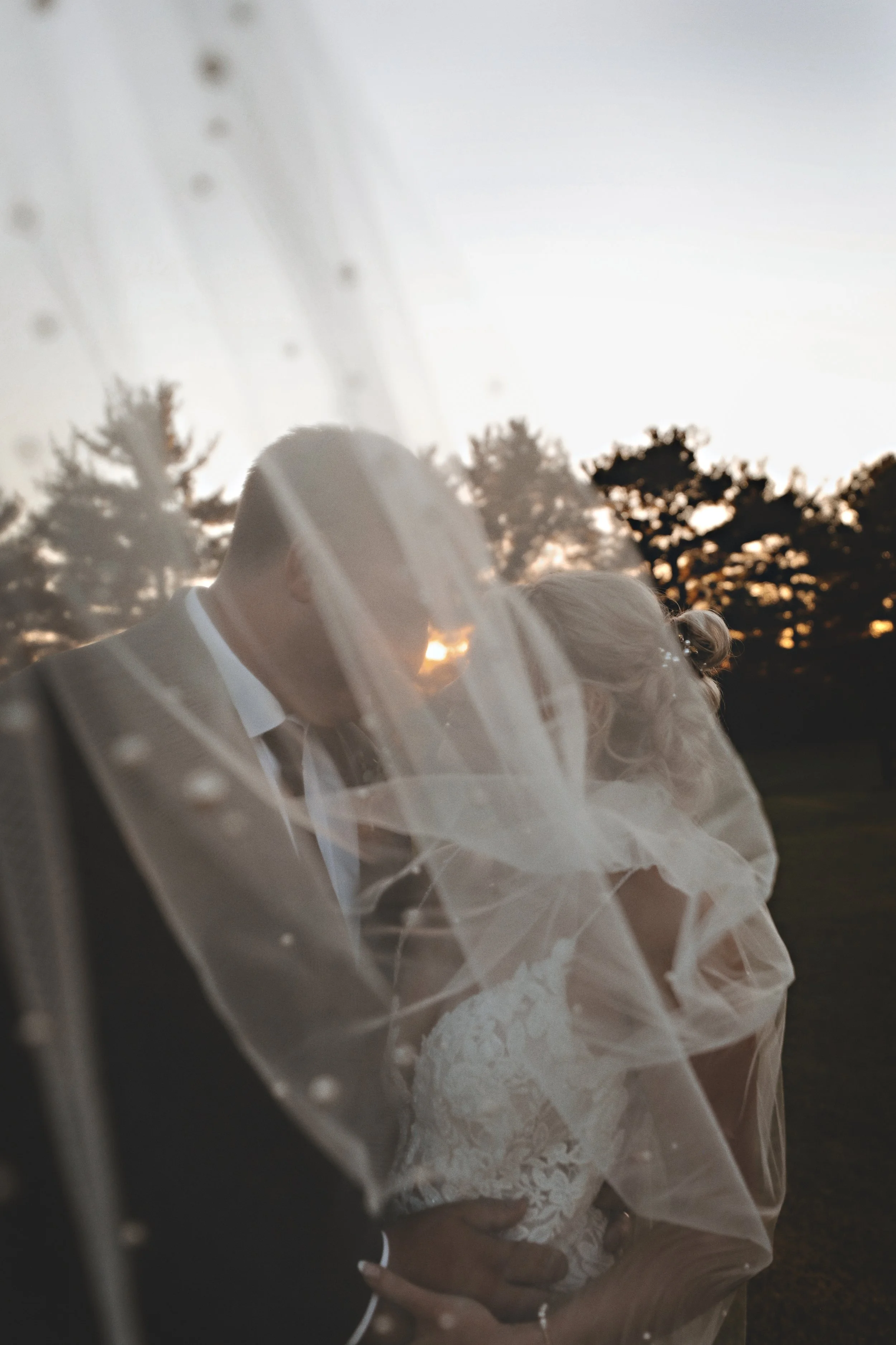 A bride and groom sharing a kiss behind a wedding veil with a sunset and trees in the background.