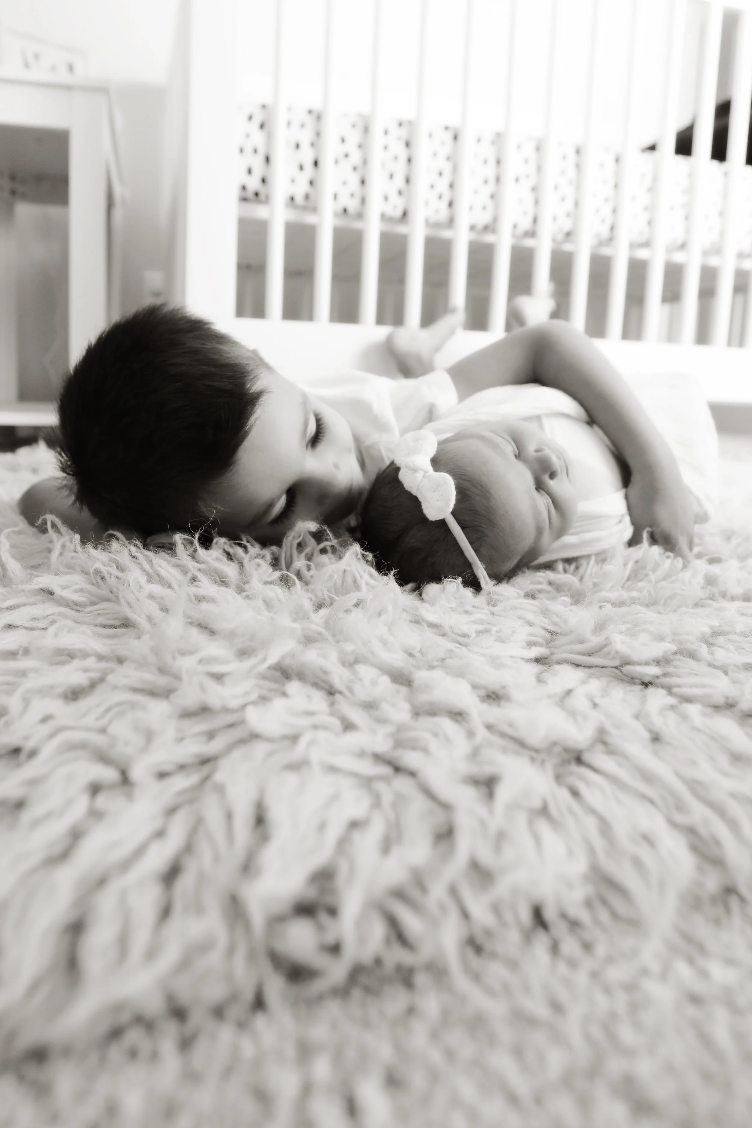 A young boy and a newborn baby laying on a soft, textured blanket in a cozy nursery, with a crib in the background.