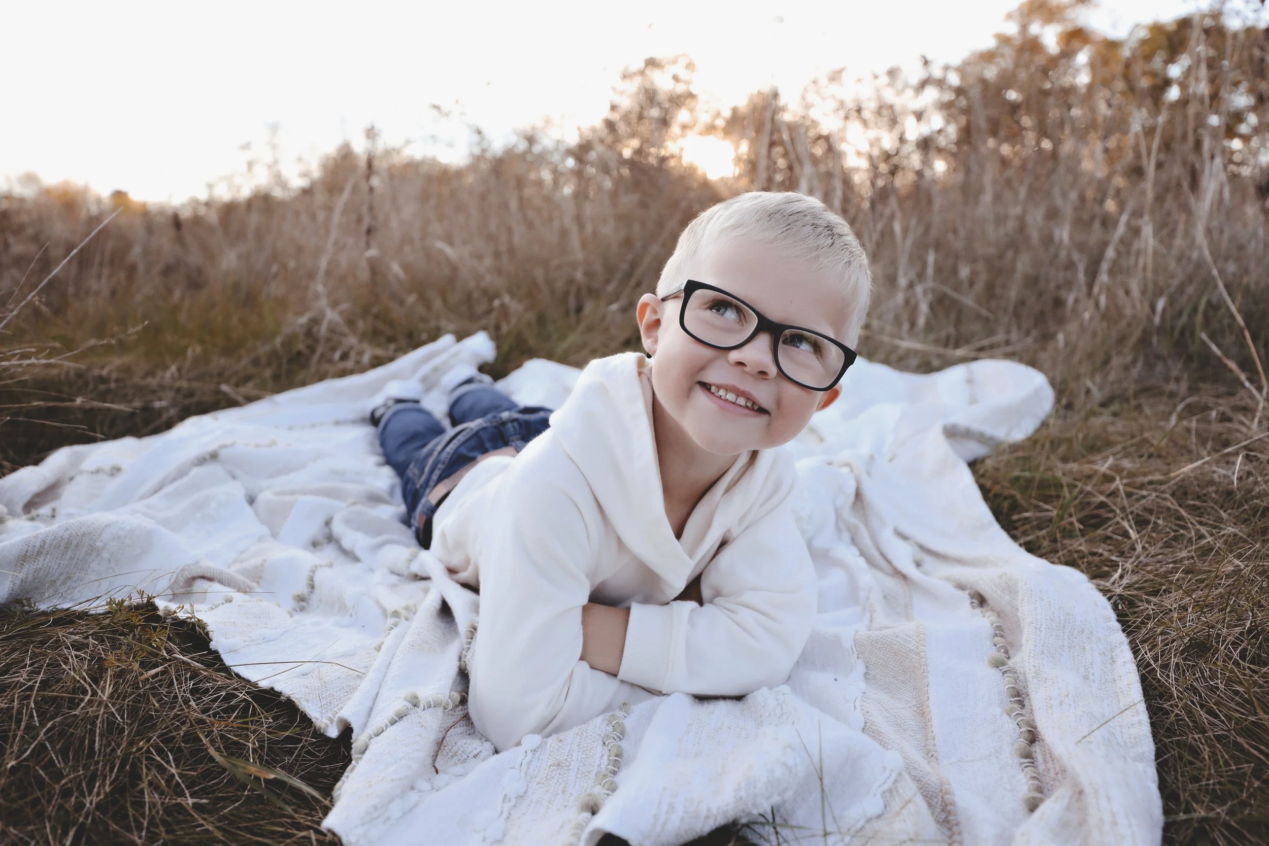 A young boy with glasses lying on his stomach on a white blanket outdoors in a field with tall, dry grass during sunset, smiling and looking up.