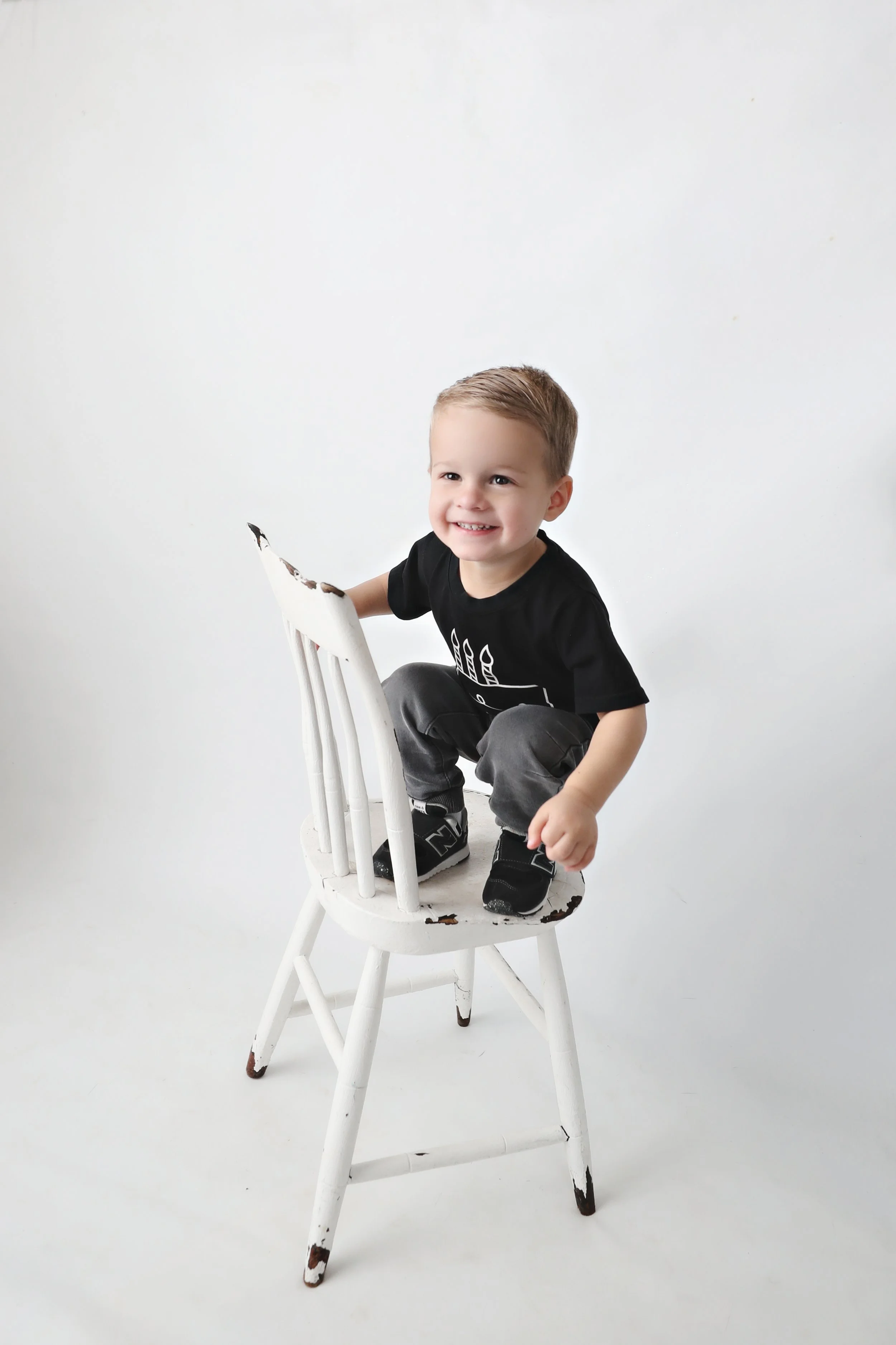 Smiling young boy crouching on a distressed white wooden chair with peeling paint, against a plain white background.