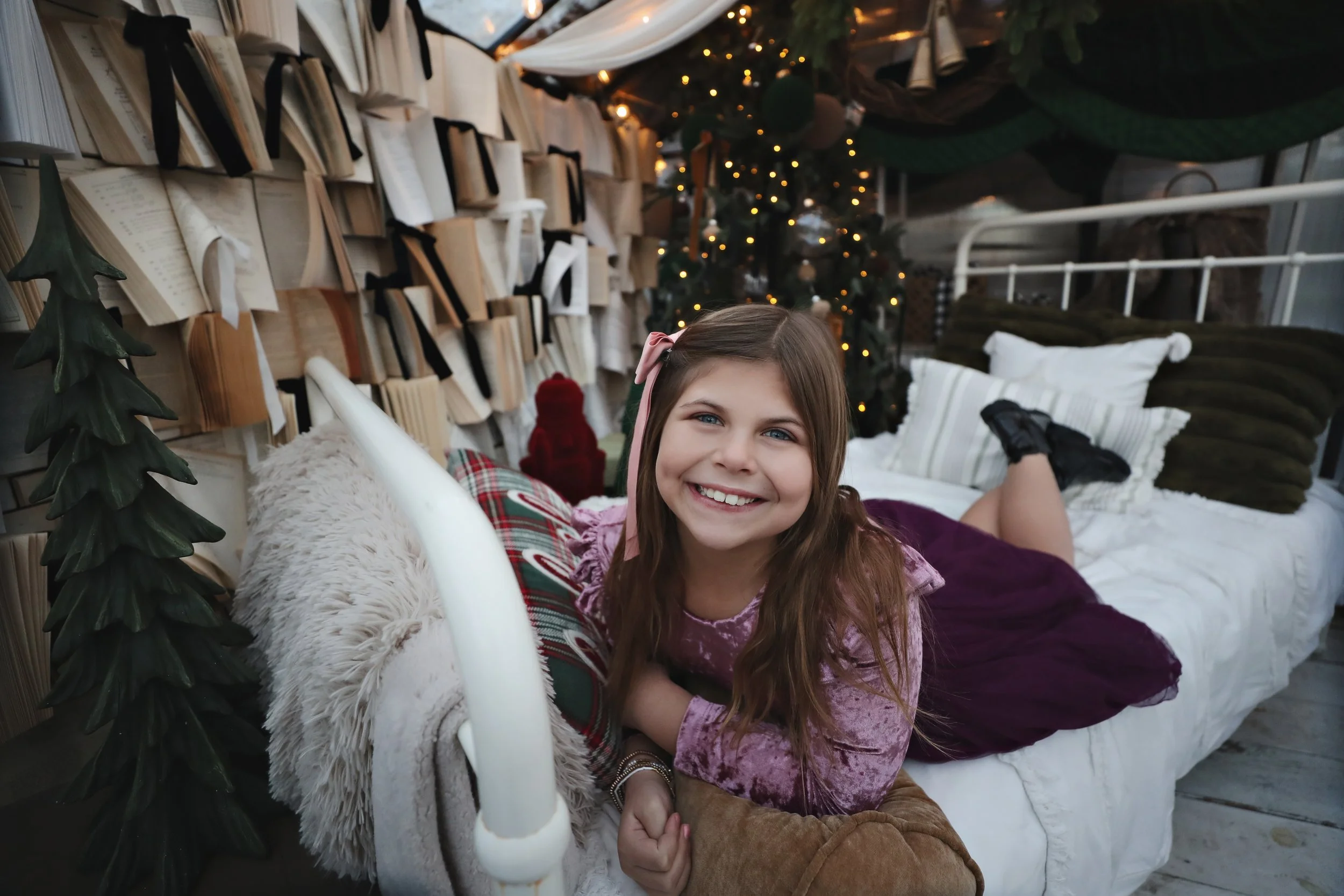A young girl lying on a holiday-themed bed, smiling at the camera. The background features a decorated Christmas tree with lights, surrounded by wrapped presents and festive pillows.