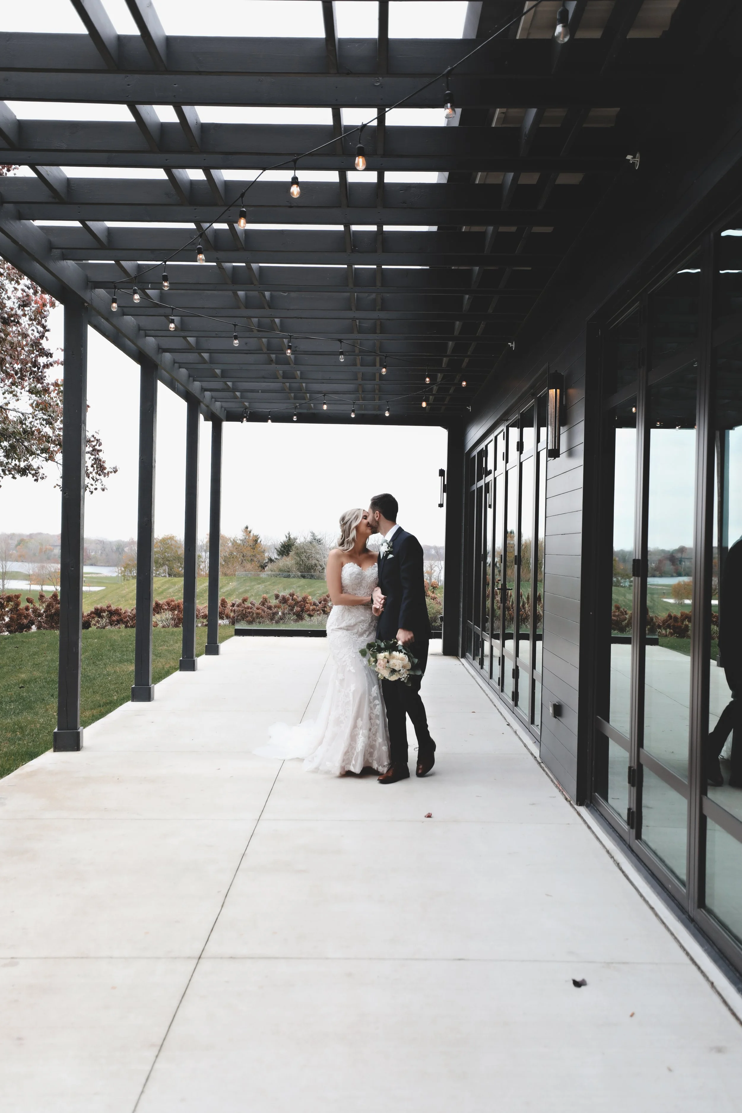 A bride and groom kiss under a covered patio on their wedding day, with outdoor scenery in the background.