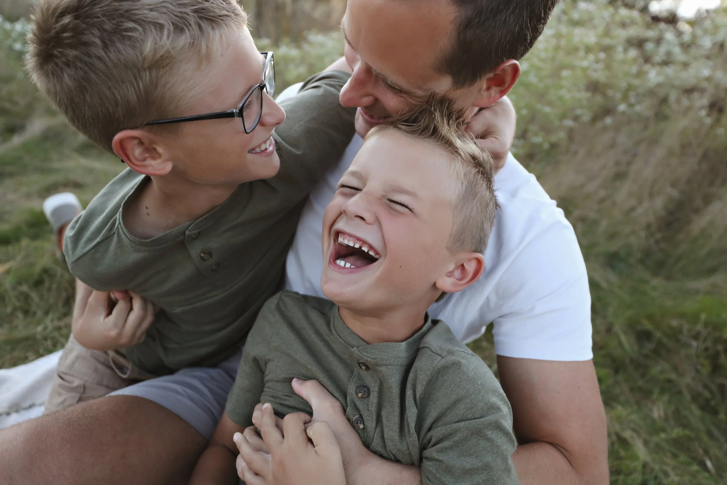 A man laughing and hugging two young boys with blonde hair outdoors in a grassy field.