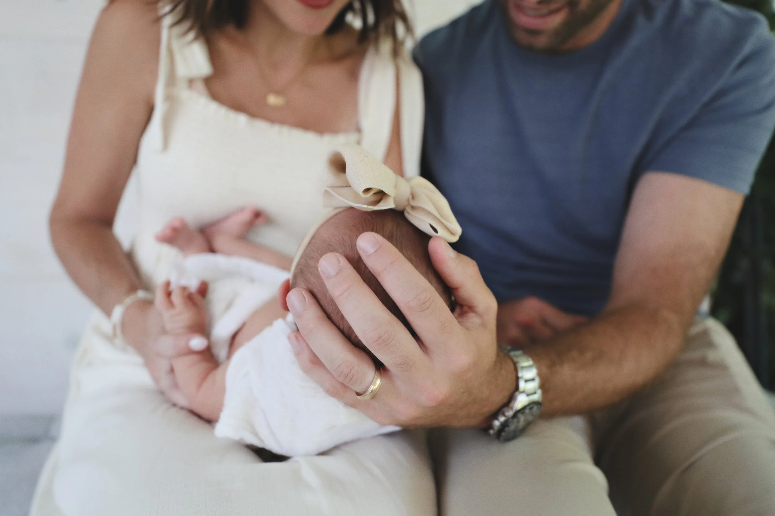 A woman and man holding a baby with a large beige bow on the baby's head.