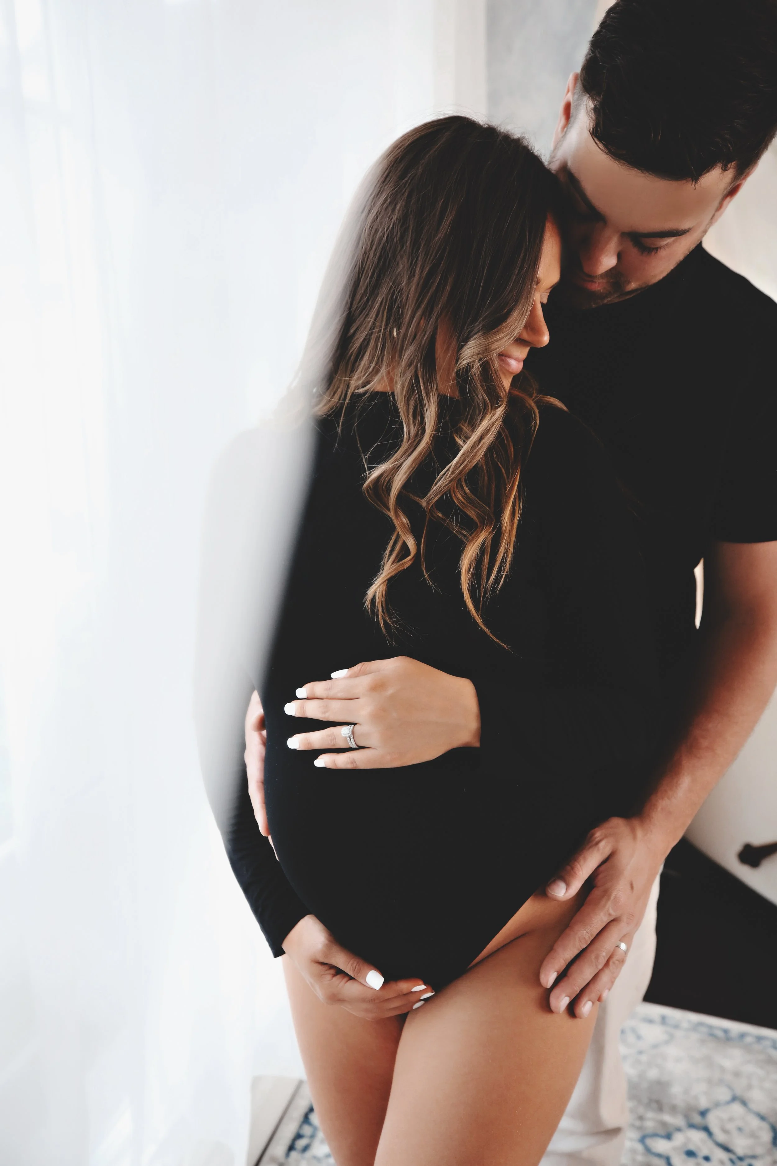 A couple embracing affectionately indoors, with the woman showing her pregnancy and wearing a ring on her left hand.