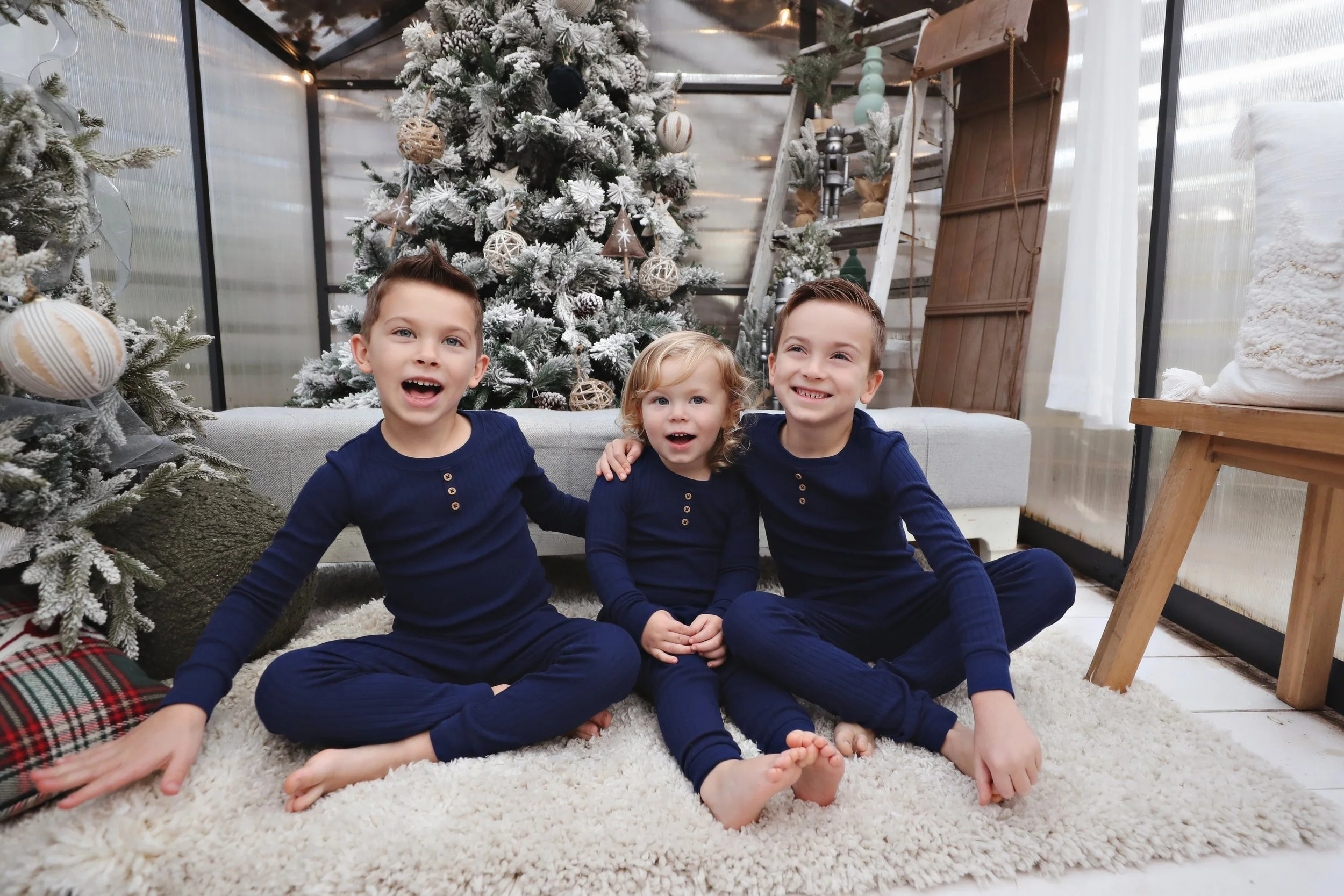 Three children sitting on a cozy white rug in front of a decorated snow-covered Christmas tree, inside a room with a glass ceiling and wooden furniture, celebrating Christmas.