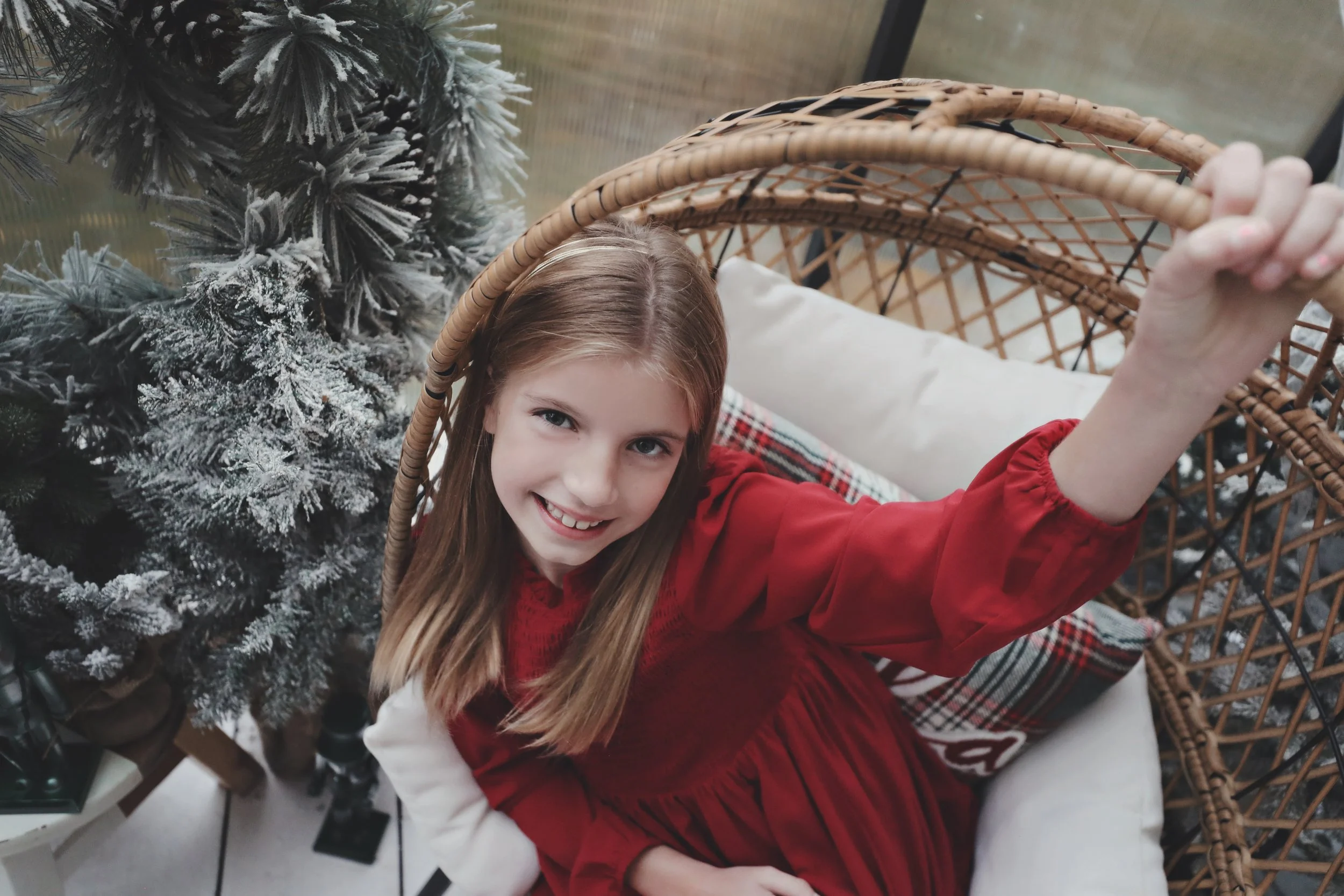 A young girl with long blonde hair smiling and reaching up inside a wicker hanging chair. There is a snow-dusted Christmas tree next to her, and she is wearing a red dress.