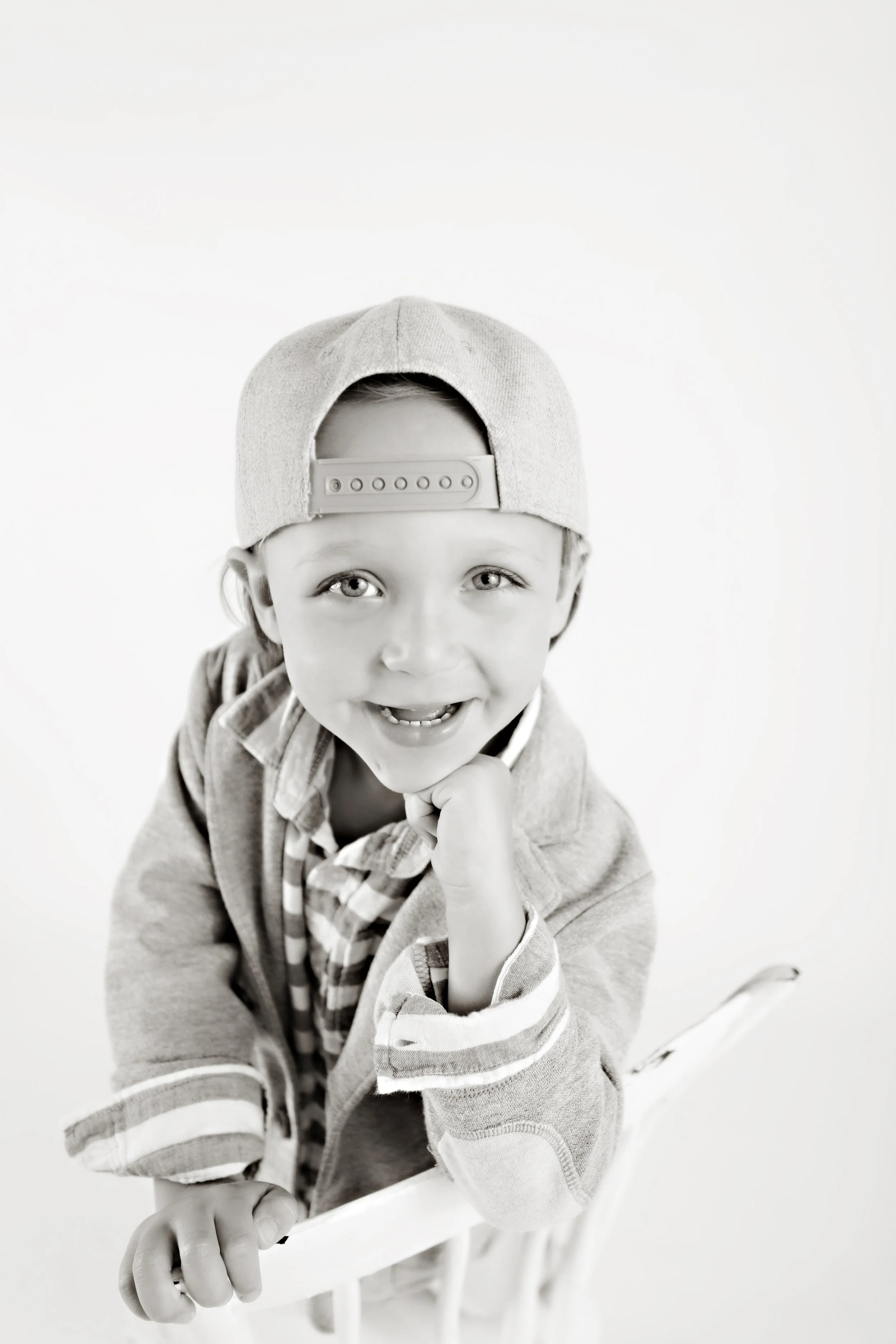 Young boy with a backward cap, smiling at the camera, resting his chin on his hand.