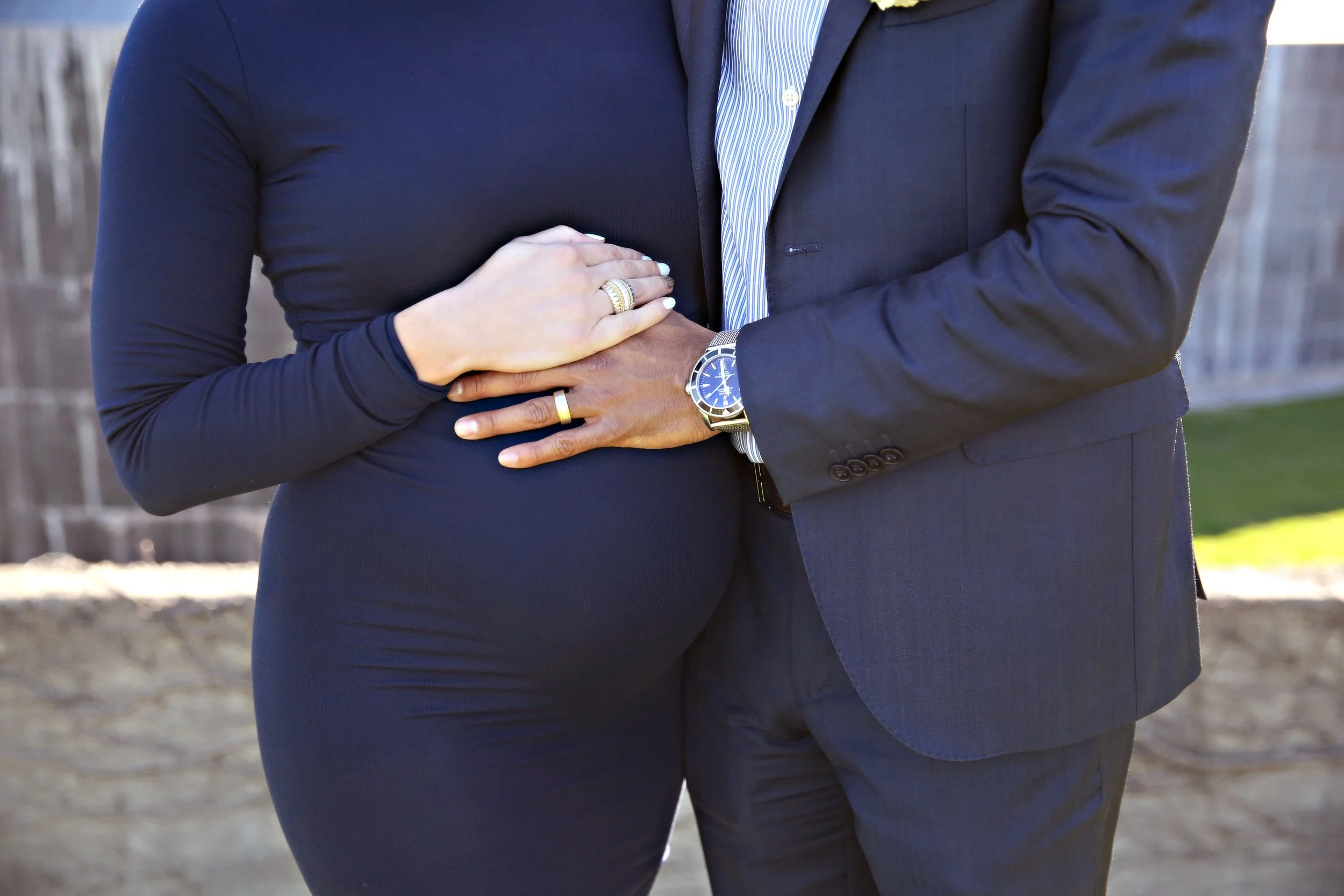 A close-up of a man's and woman's hands on her waist, showing rings and a watch, outdoors in daylight.