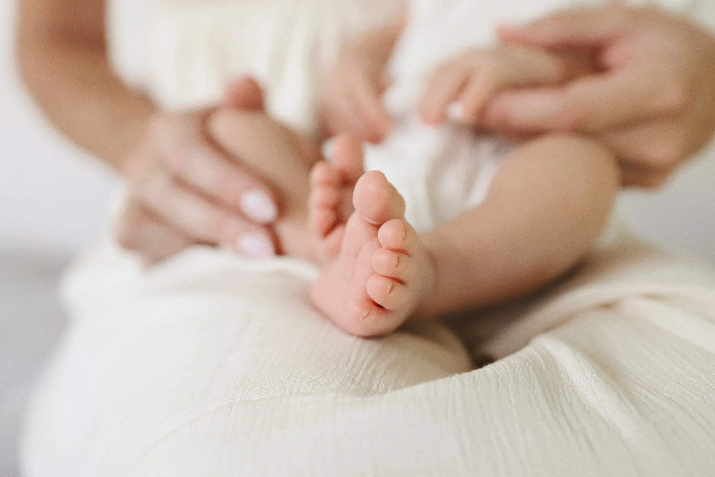 Close-up of a baby's toes being gently held by adult hands, with the baby lying on a soft, light-colored fabric.