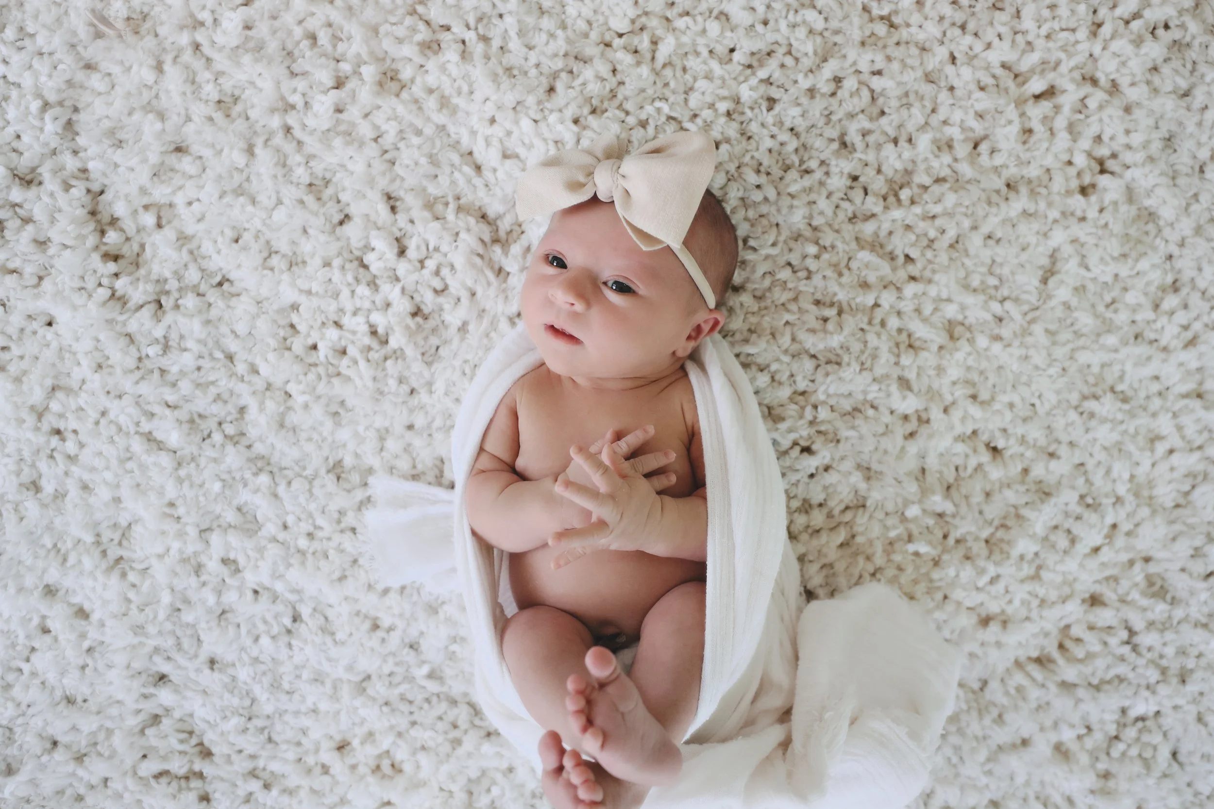 A baby lying on a fluffy white blanket, wearing a beige headband with a large bow and a white cloth draped around their shoulders.