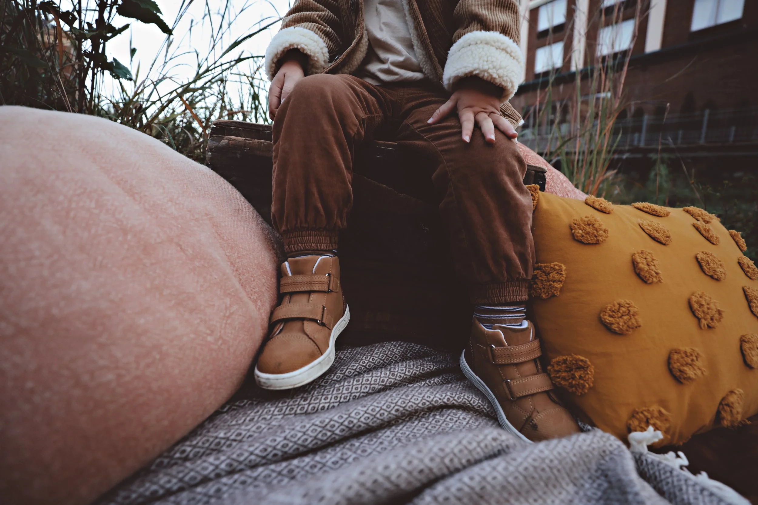 Child sitting outdoors on a bed of cushions with one hand resting on their thigh, wearing brown pants, tan shoes, and striped socks, with a background of plants and a building.