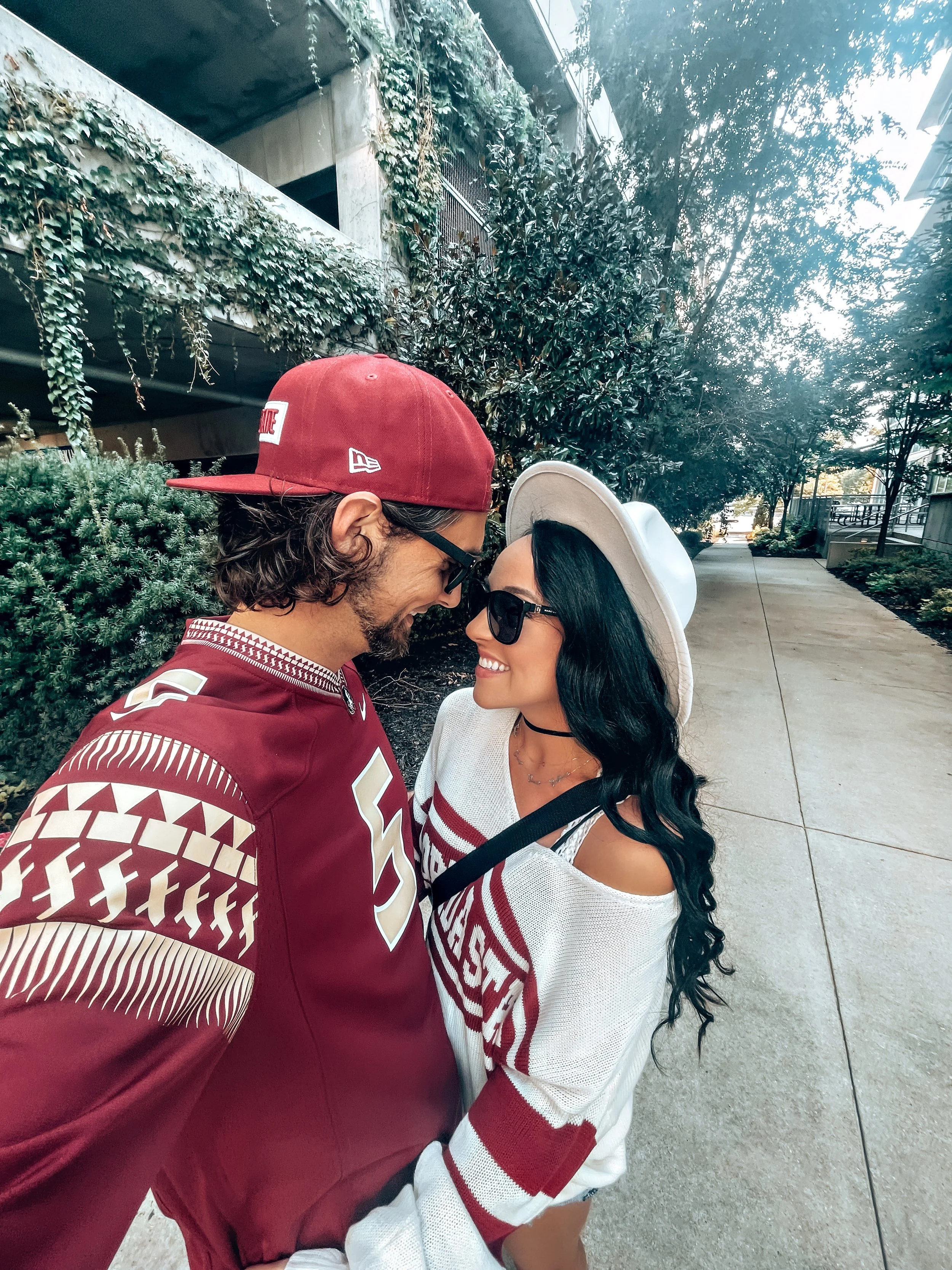 A man and woman smiling and touching foreheads on an outdoor sidewalk, with trees and buildings in the background. The man is wearing a red cap, sunglasses, and a maroon sports jersey. The woman is wearing a white hat, sunglasses, and a white sweater