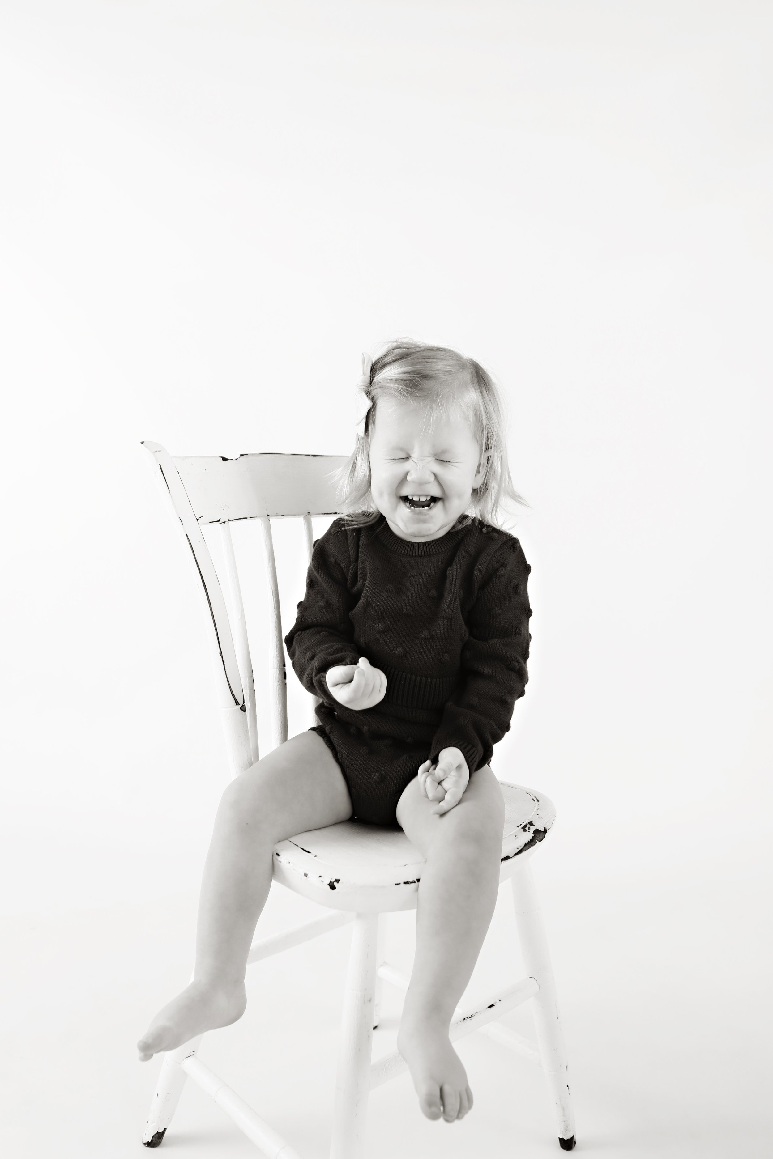 A black-and-white photo of a young girl sitting on a white wooden chair, laughing with her eyes closed, wearing a dark sweater and shorts.