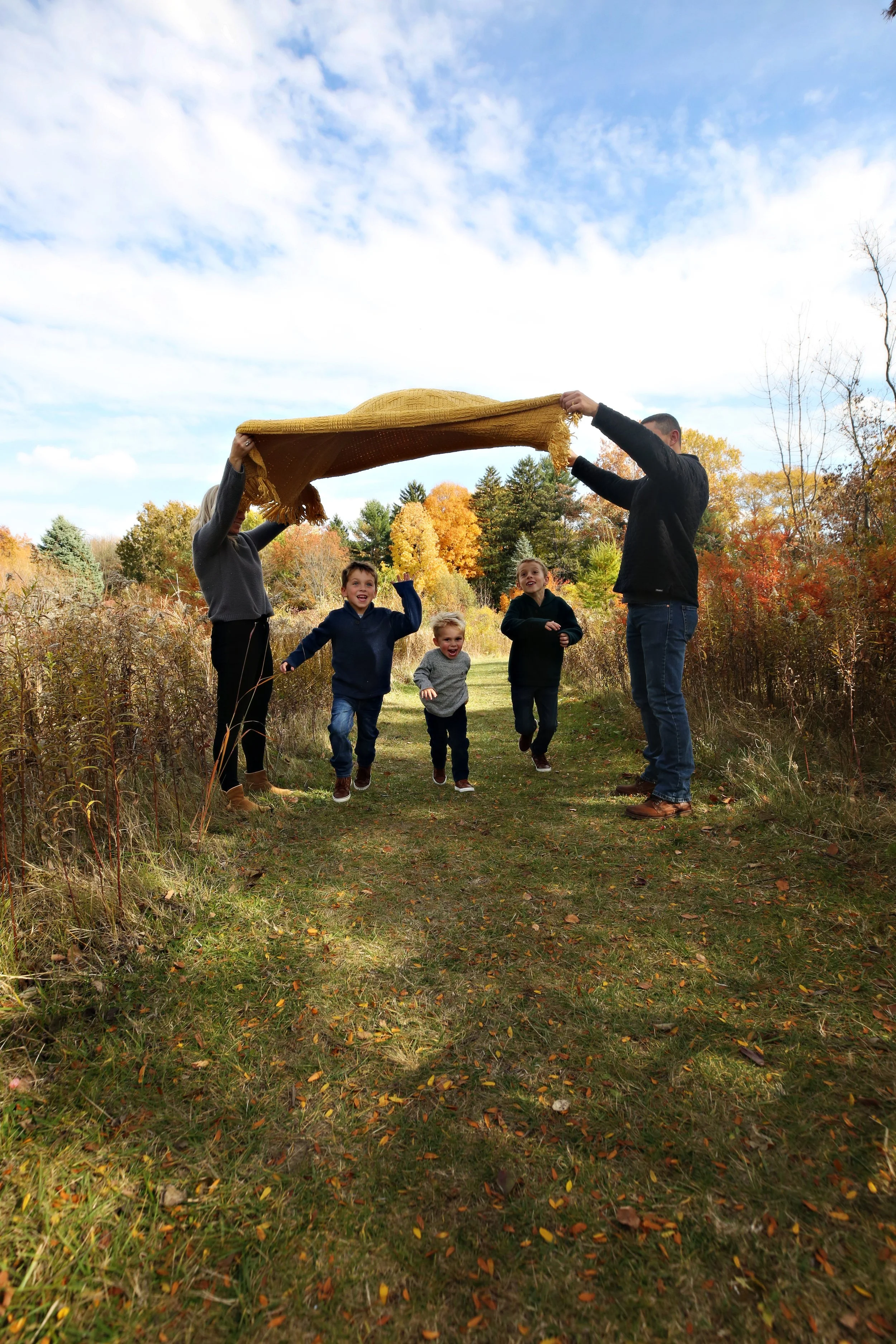 A family of five, including two adults and three children, playing outdoors in an autumn setting, running under a yellow blanket held overhead by the adults.