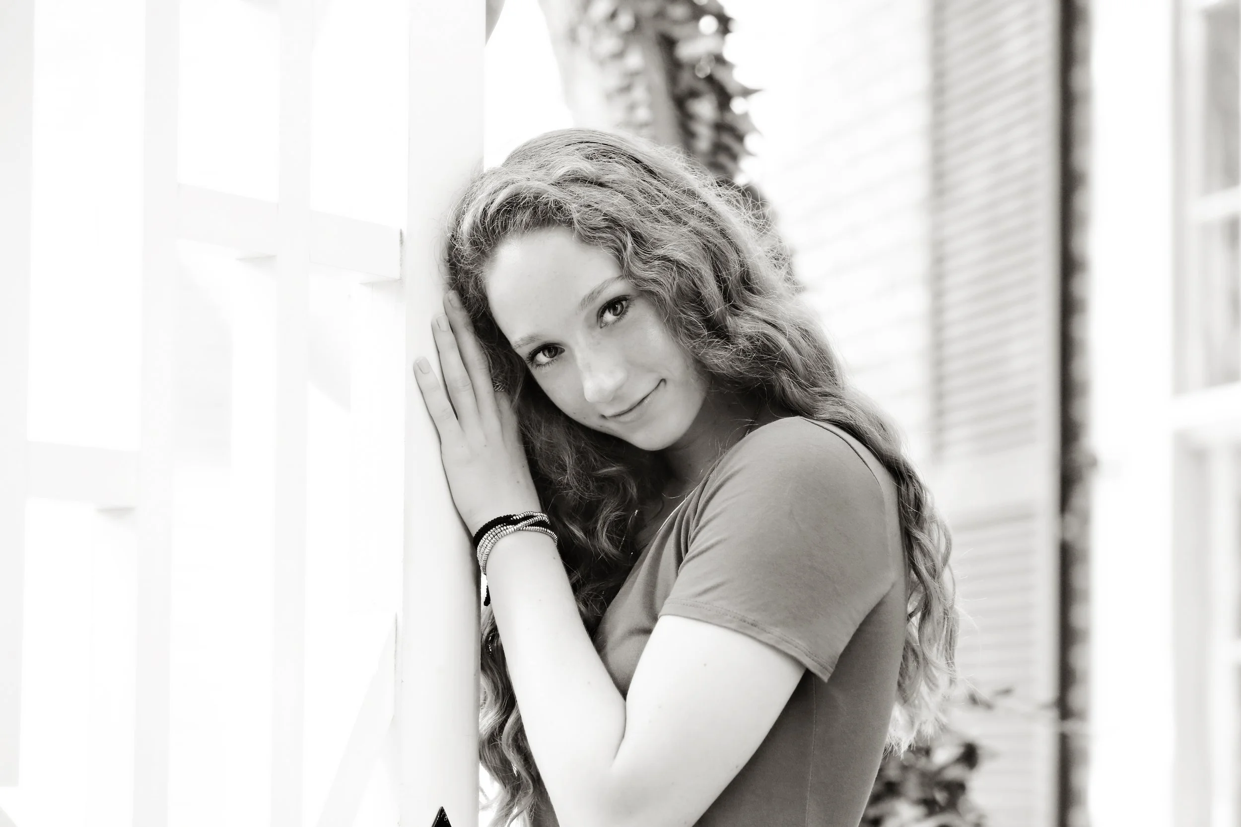 Black and white photo of a young woman with long curly hair, leaning against a wall with her hands near her face, smiling softly.