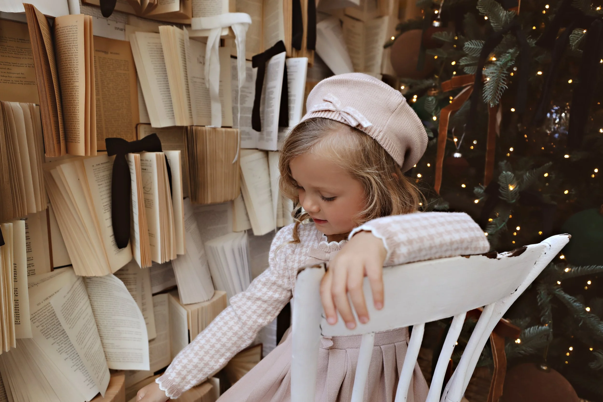 A young girl with curly hair wearing a beige beret and a pink dress, sitting on a white chair, reaching towards an open book on a shelf of books, decorated with black ribbons, near a decorated Christmas tree with lights and ribbons.
