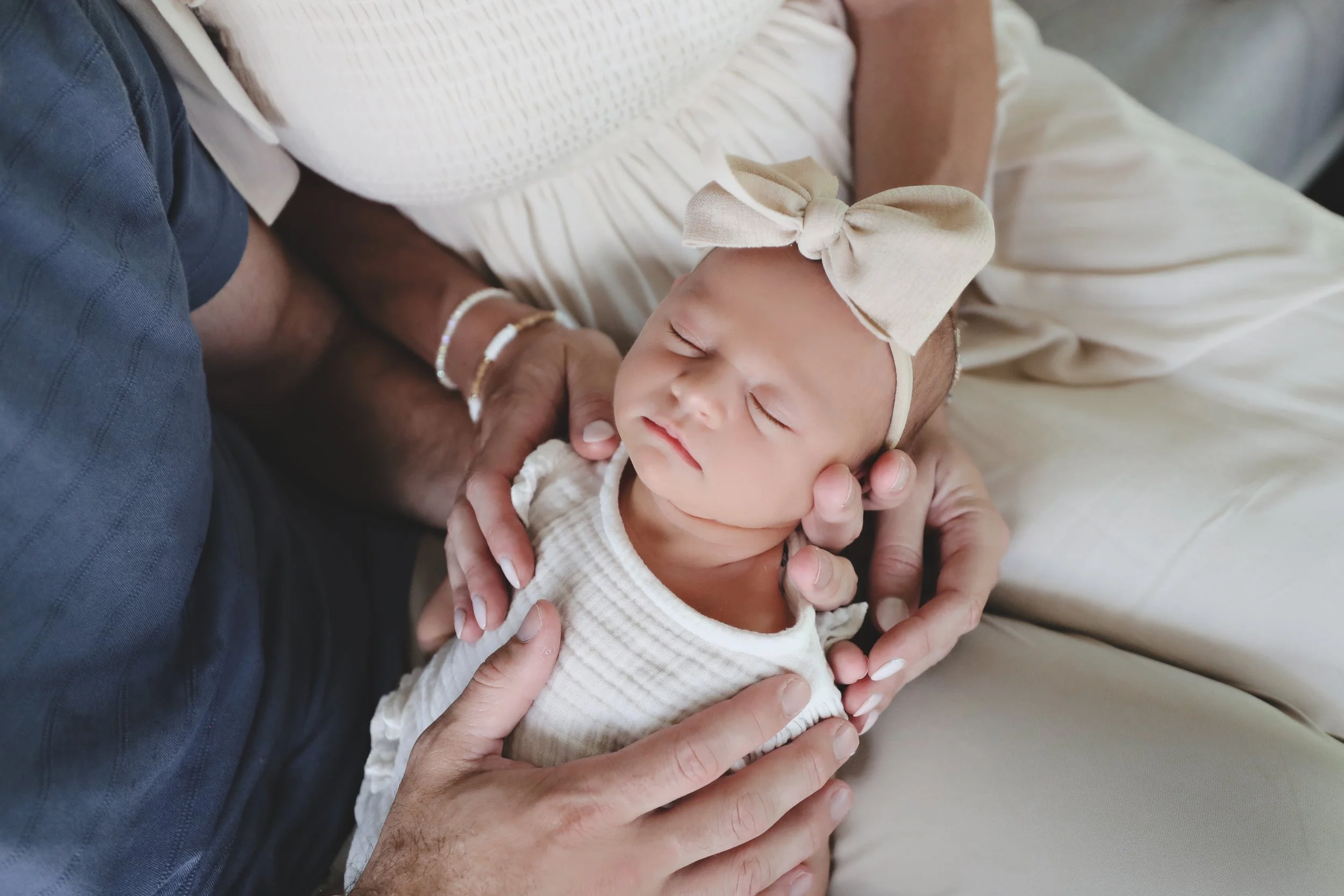 Close-up of a sleeping infant girl wearing a beige headband with a large bow, held gently by an adult with multiple bracelets, surrounded by loving hands.