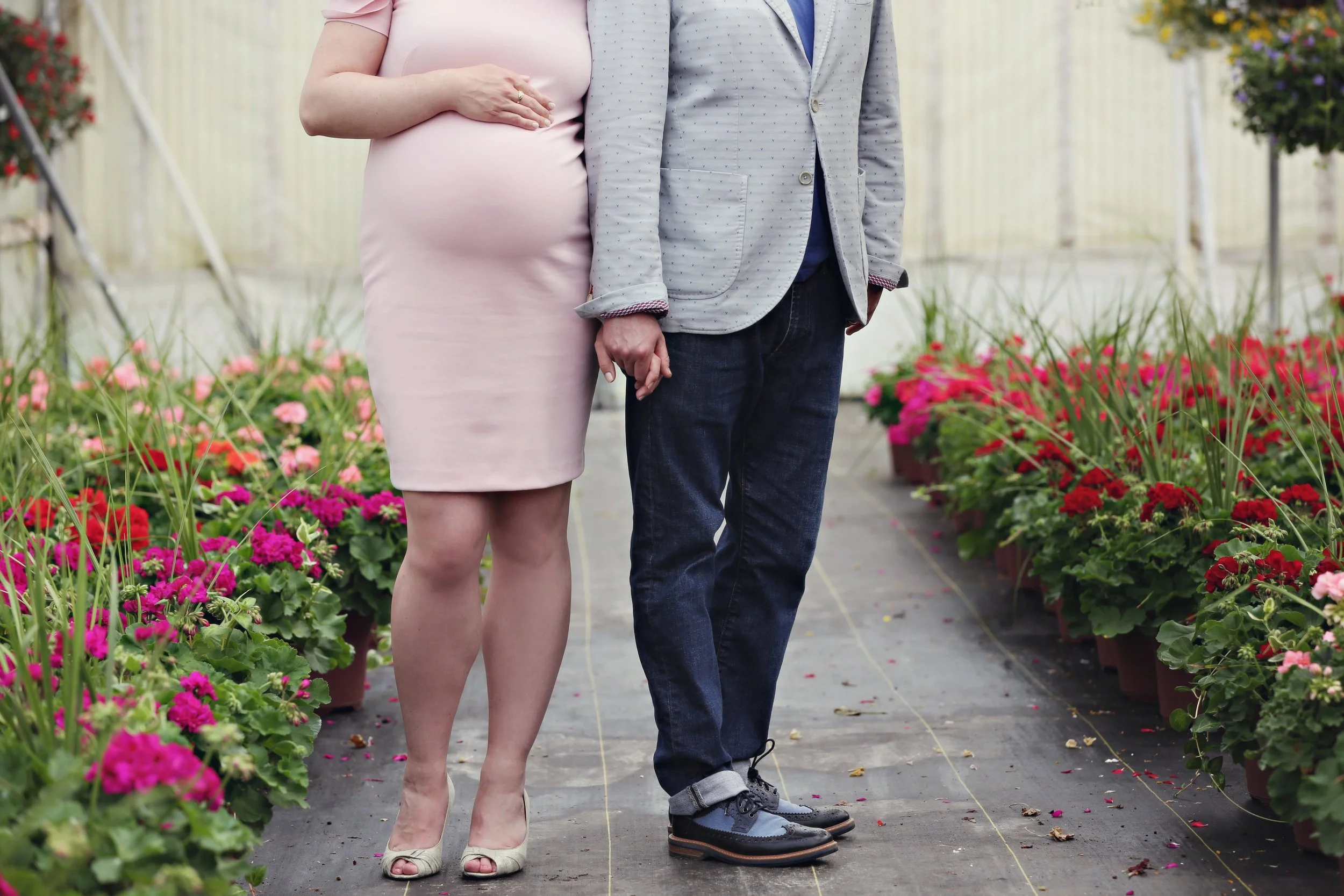 A pregnant woman in a light pink dress holding hands with a man in a gray blazer and dark jeans, standing in a greenhouse surrounded by colorful flowers.