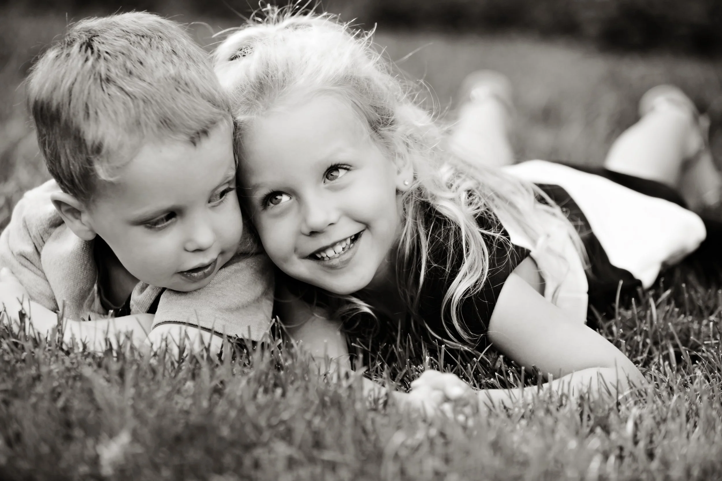 Two children, a boy and a girl, lying on grass, smiling and looking at each other, captured in black and white.