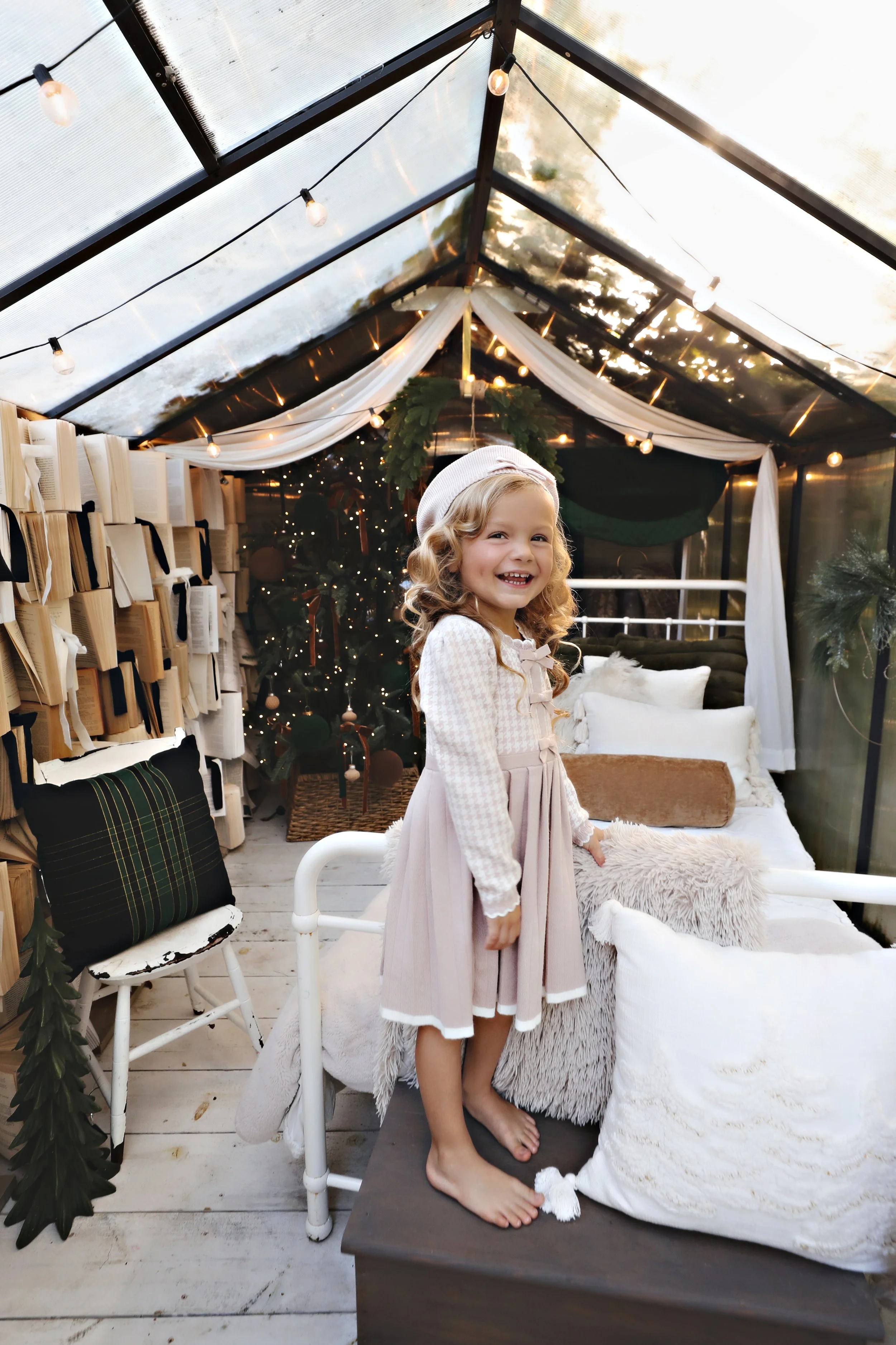 A smiling young girl with curly blonde hair, wearing a white and beige dress and a white hat, standing on a cushioned bed in a decorated indoor space with Christmas trees, fairy lights, and cozy pillows.