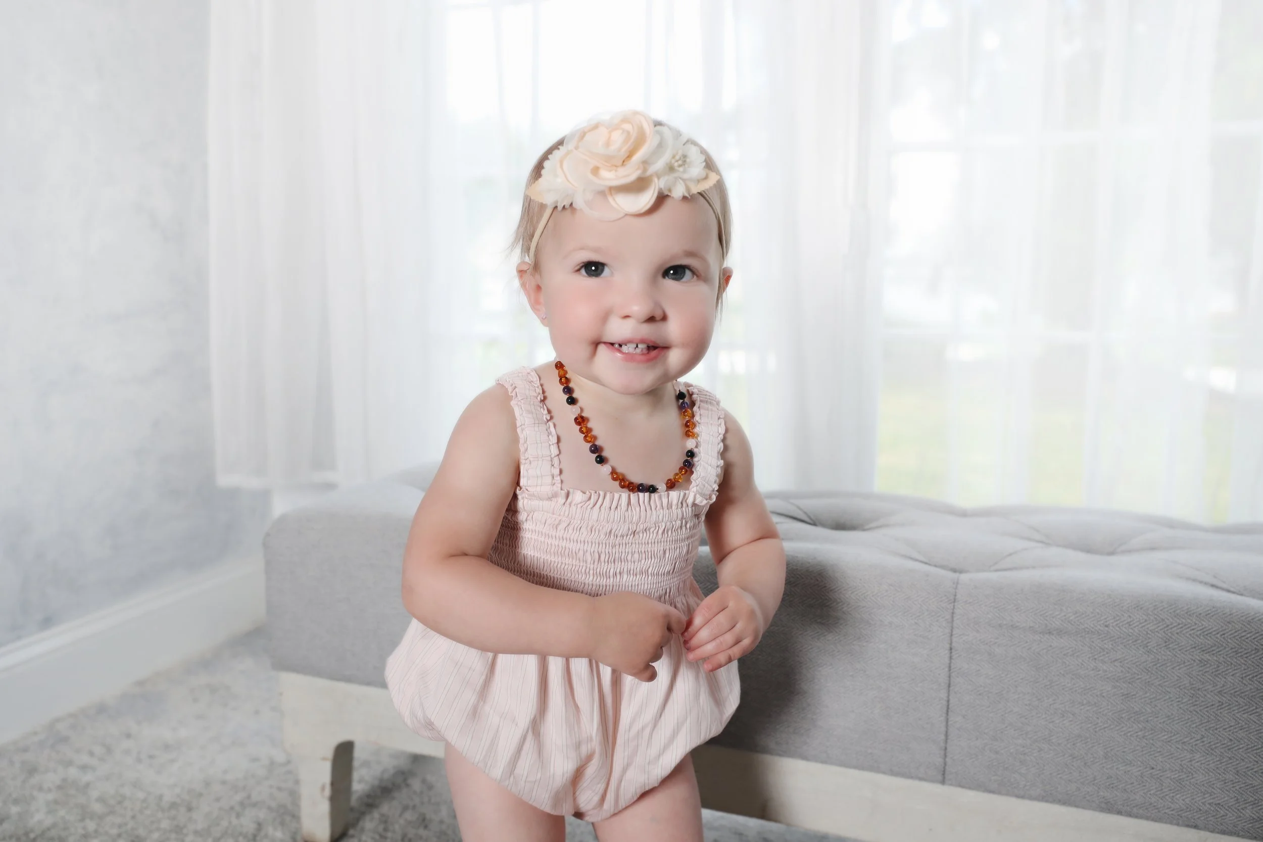 A young girl with a large flower headband and a beaded necklace, smiling and standing in a bright room with white curtains and a gray ottoman.