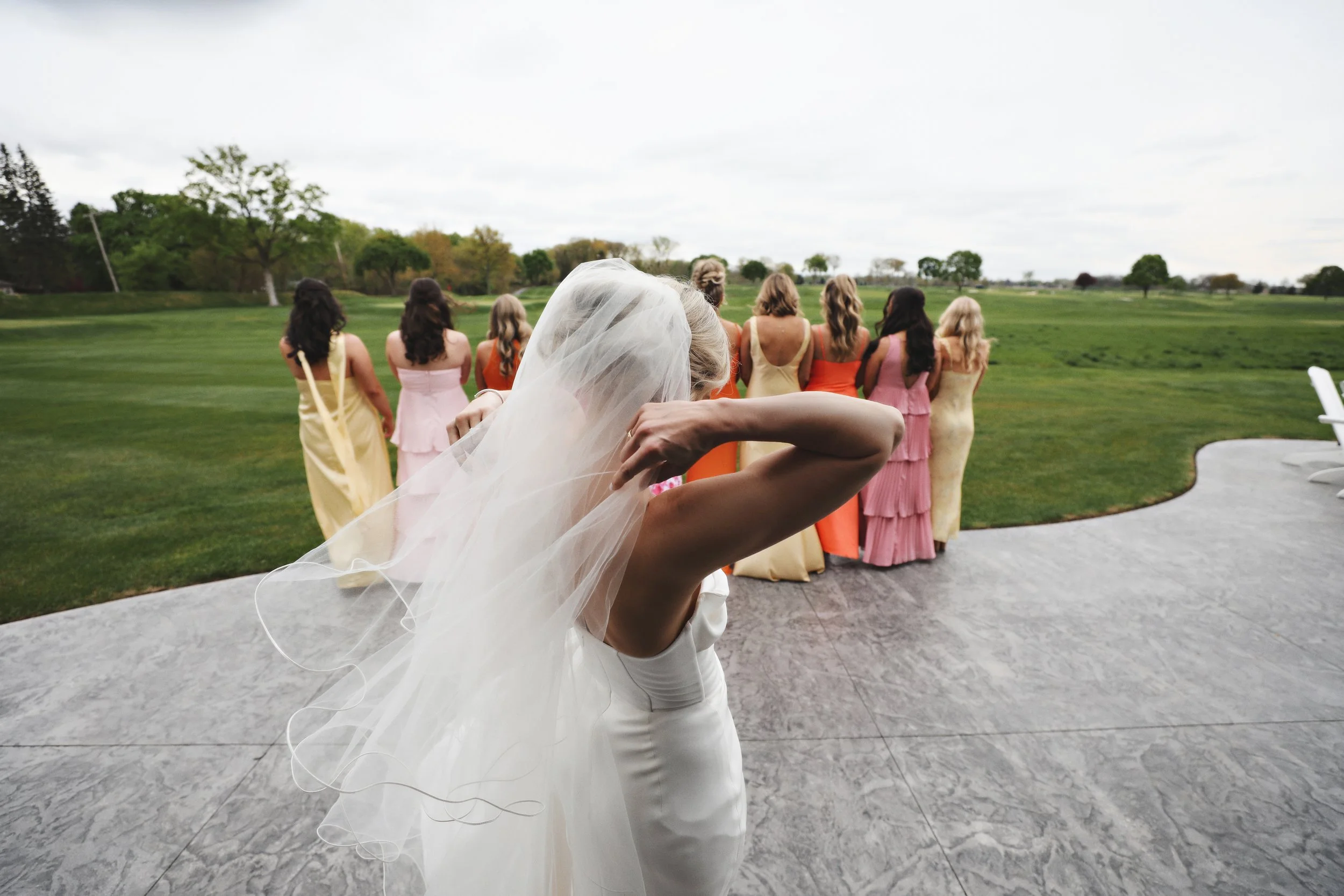 A bride in a white wedding dress and veil is adjusting her hair on a patio overlooking a grassy field where a group of women in colorful dresses stand in a line.