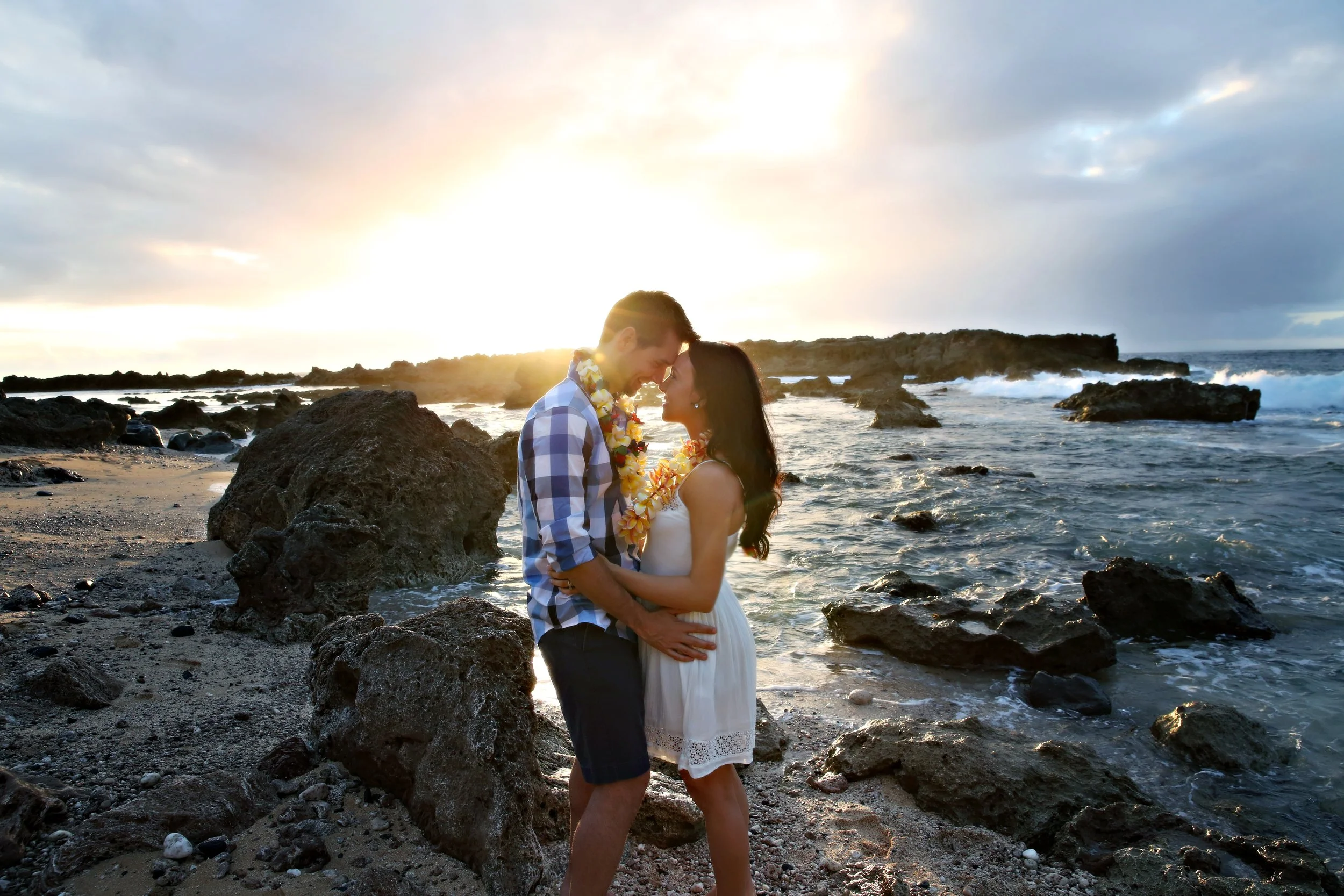 A couple stands close together on a rocky beach at sunset, facing each other with foreheads touching, while wearing flower leis. The man wears a plaid shirt and shorts, and the woman wears a white dress. The sky is partly cloudy with the sun setting 
