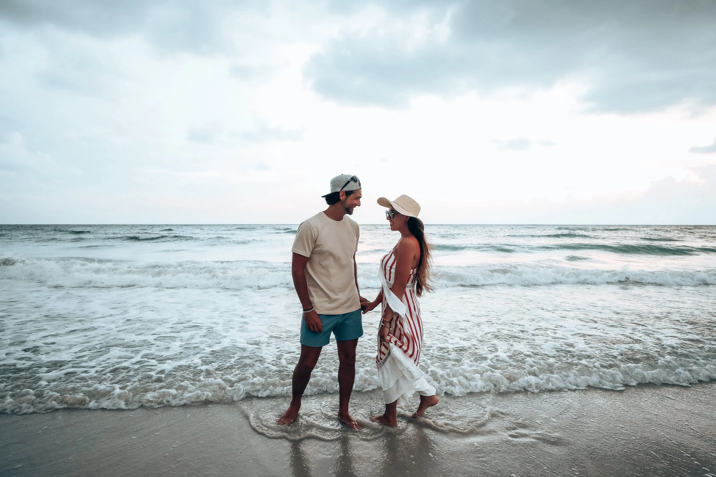 A man and woman holding hands on the beach with waves in the background, the woman wearing a sun hat and striped dress, the man wearing a gray cap and casual clothes.