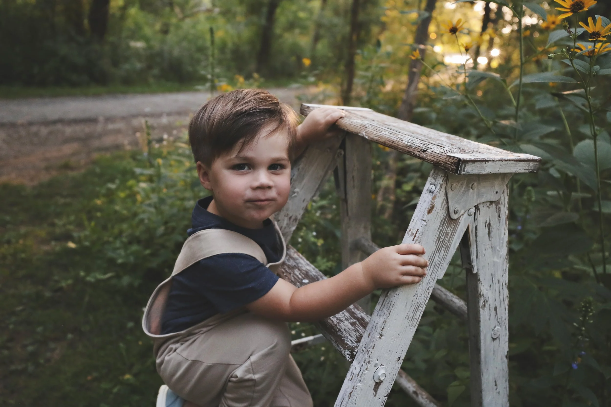 A young boy with brown hair and a serious expression, climbing a weathered wooden ladder outdoors in a lush, green forested area during sunset.