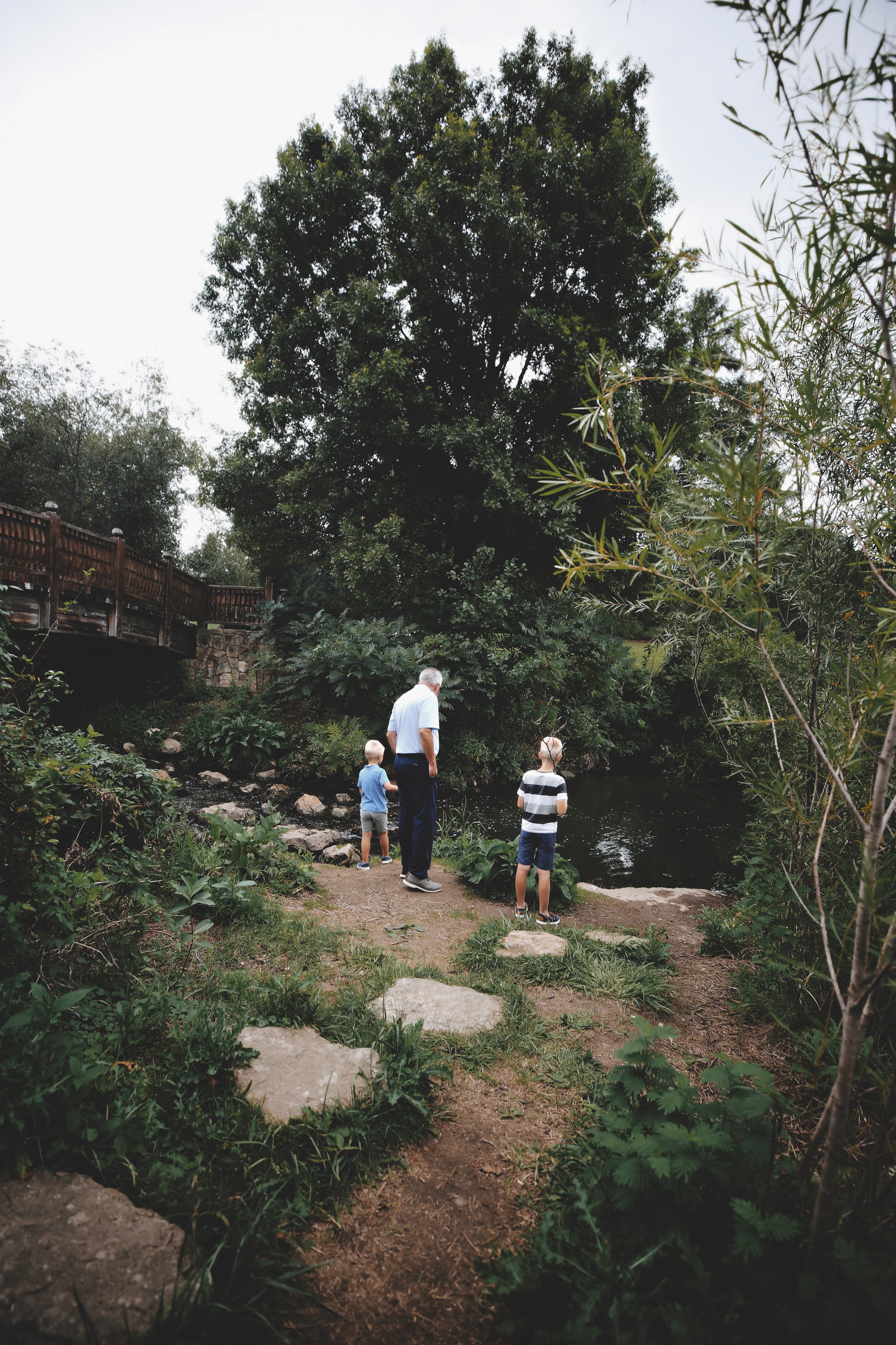 Three children and an adult stand near a river, surrounded by trees and greenery, along a wooded path with a wooden bridge in the background.