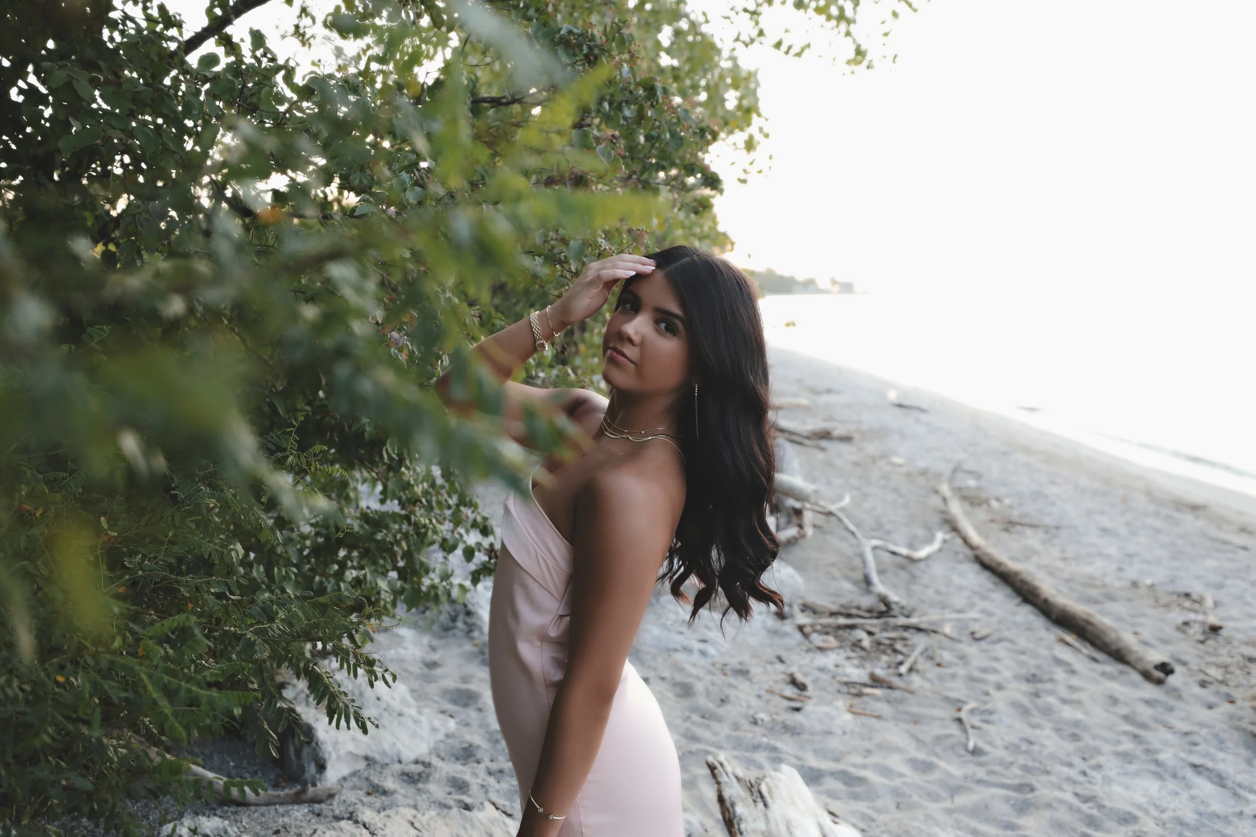 A woman in a silky pink dress standing on a beach, partially obscured by green foliage, with the ocean and sky in the background.